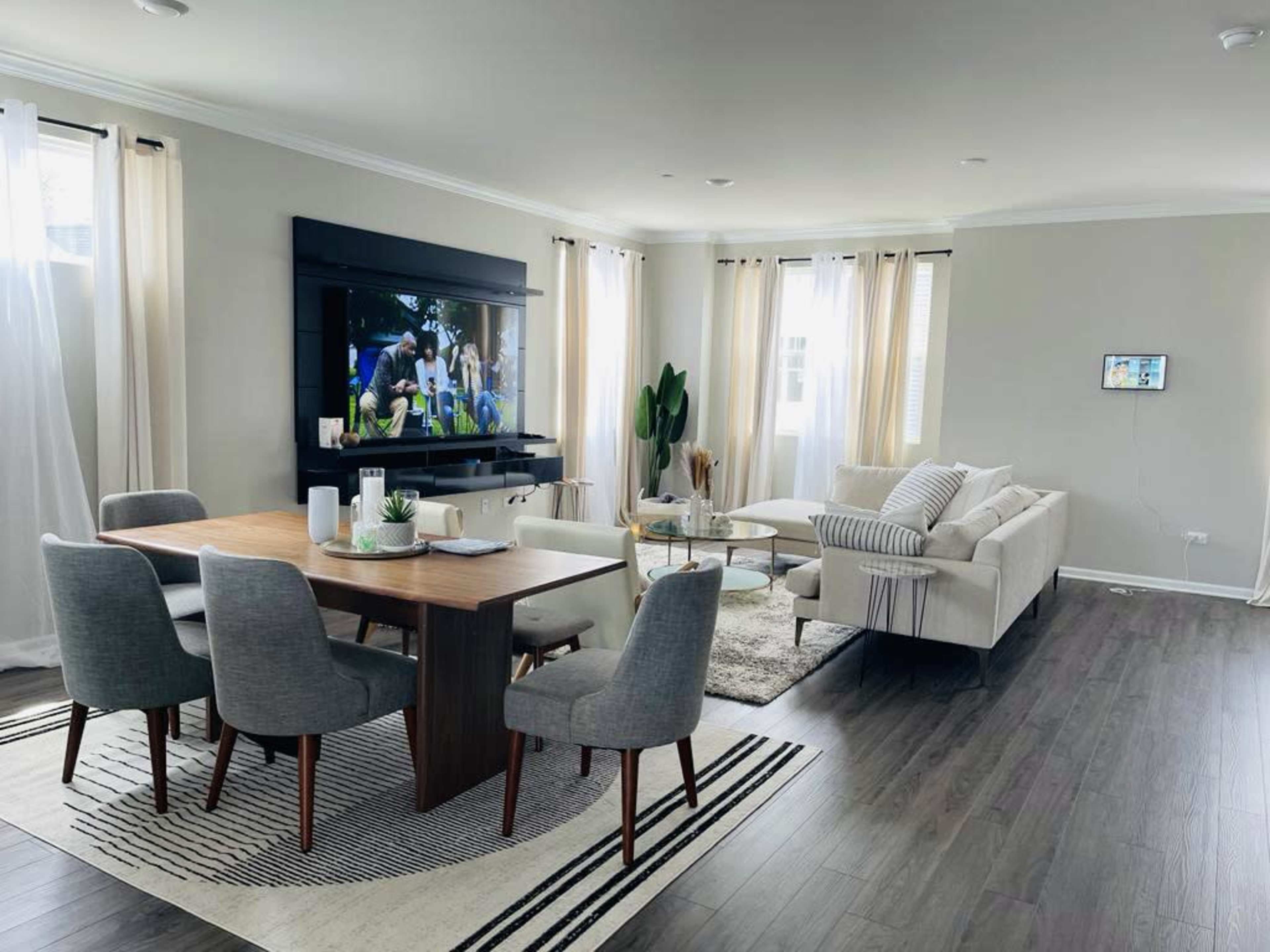 The image shows a modern living and dining area featuring a large television on the wall, a dining table with six chairs, and a light-colored sectional sofa.