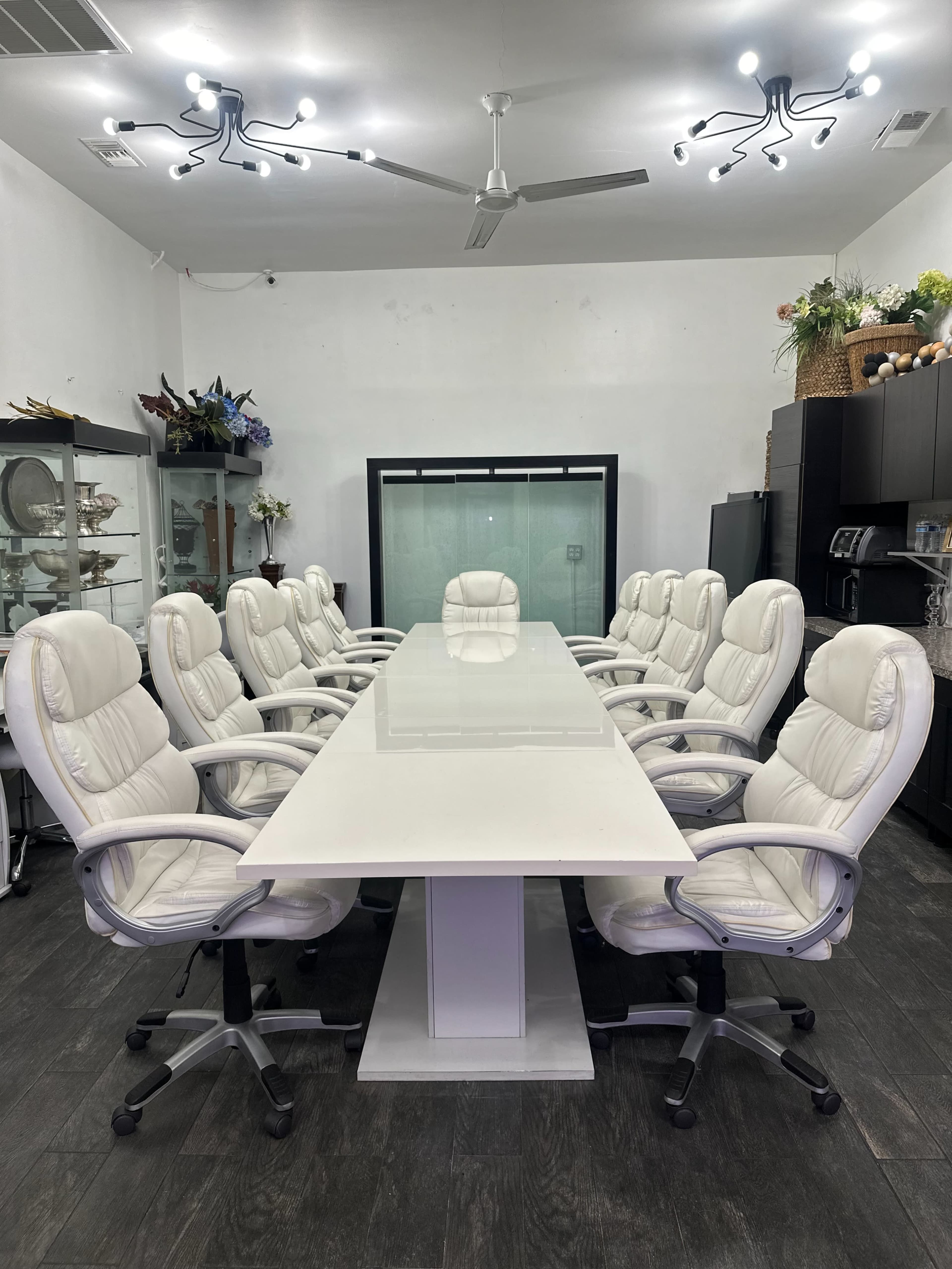 A large, modern conference room features a white rectangular table surrounded by twelve white office chairs.