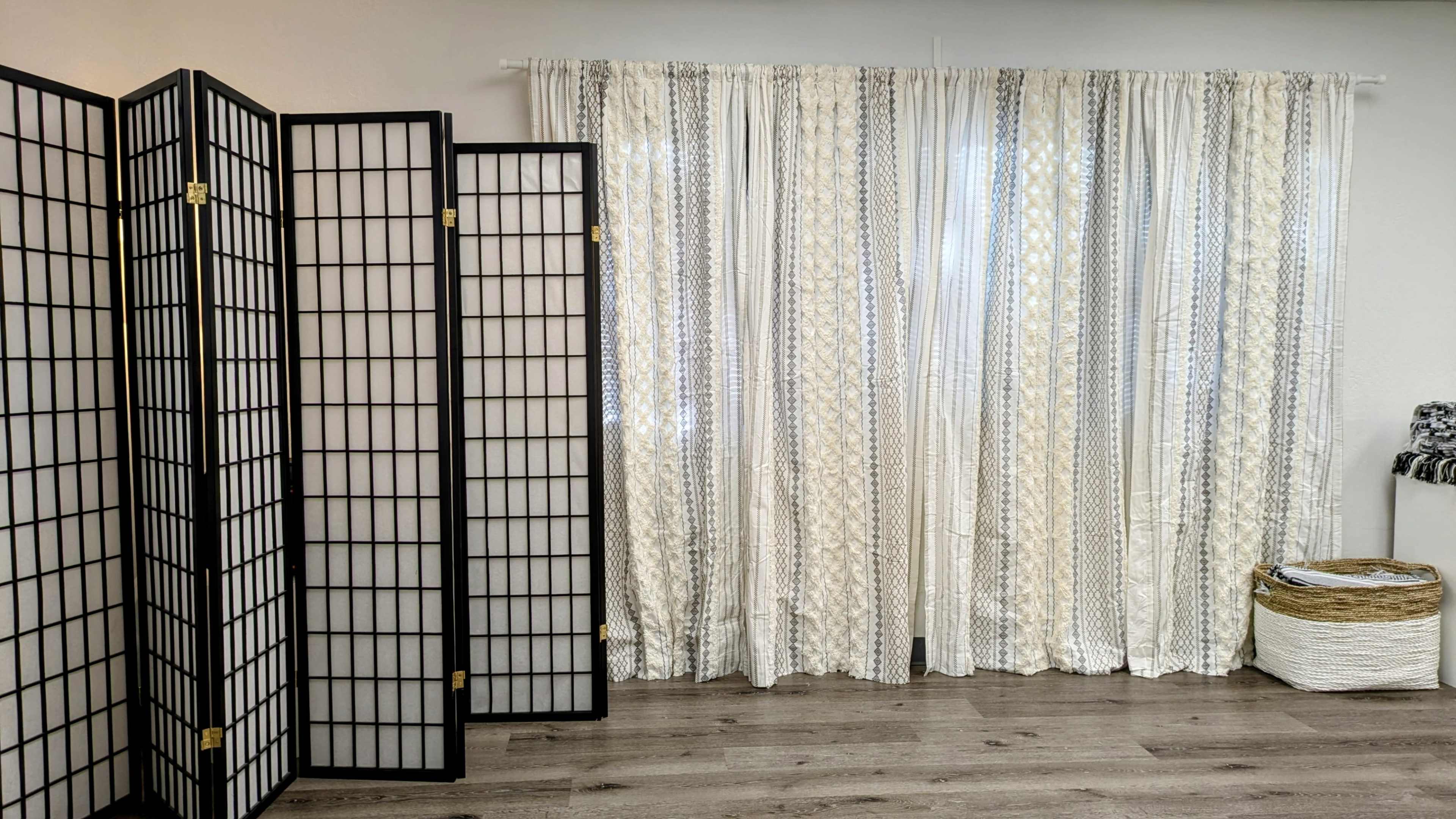 The image shows a calm interior space featuring three black folding screens, textured white curtains, and a woven basket on the floor.