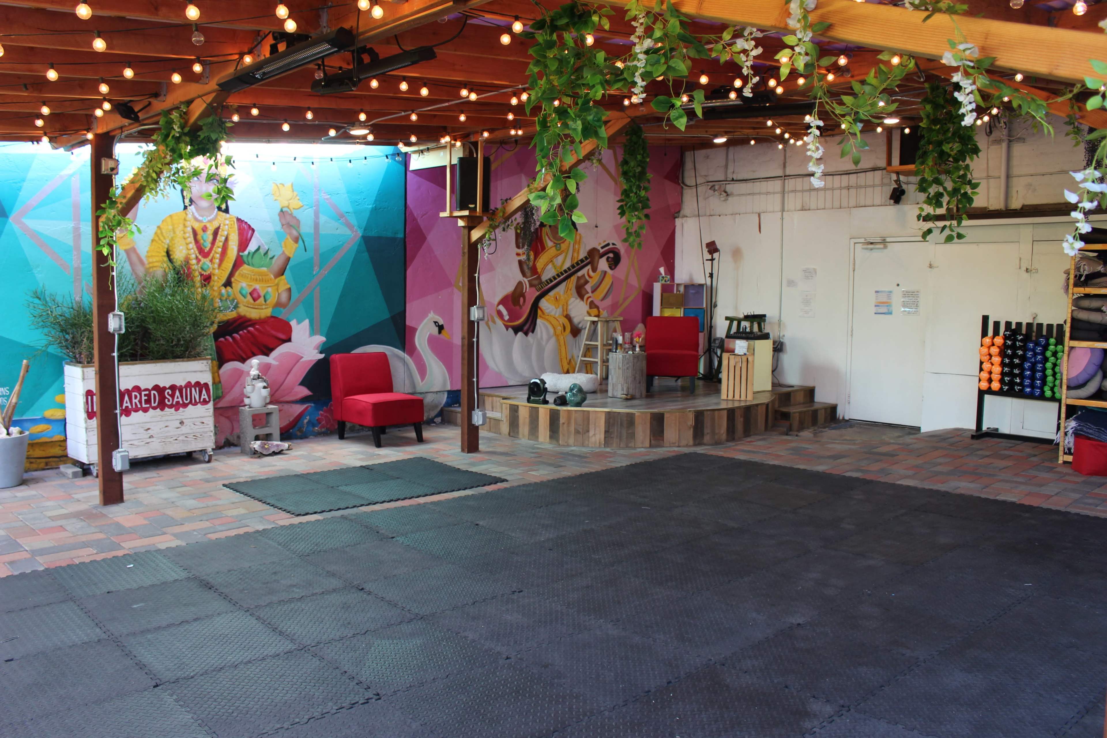 A colorful outdoor space with a wooden deck featuring a small stage, red chairs, and vibrant murals on the walls, surrounded by string lights and potted plants.
