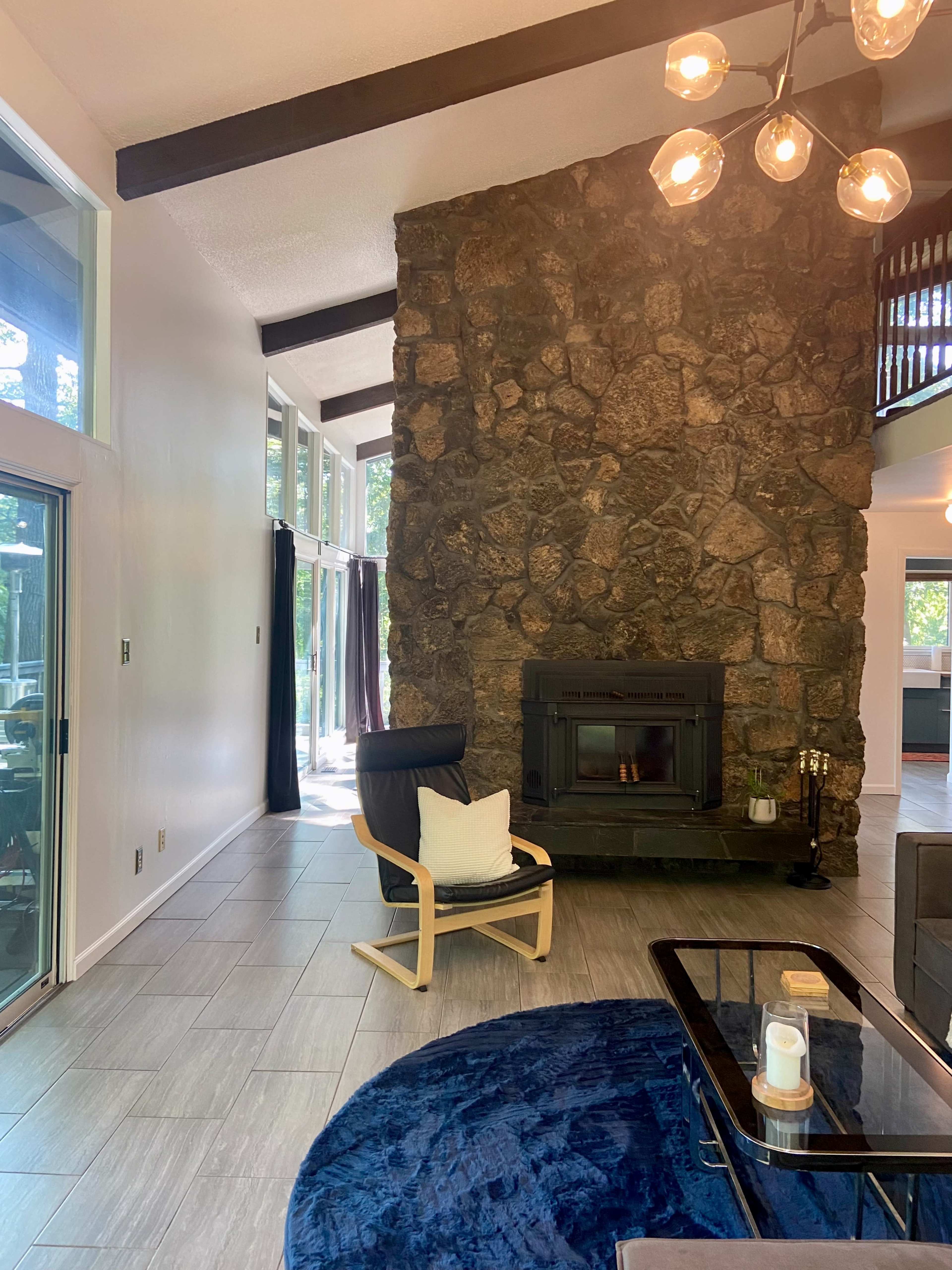 The image shows a spacious living room featuring a stone wall with a fireplace, large windows, and modern furniture including a chair and a coffee table.