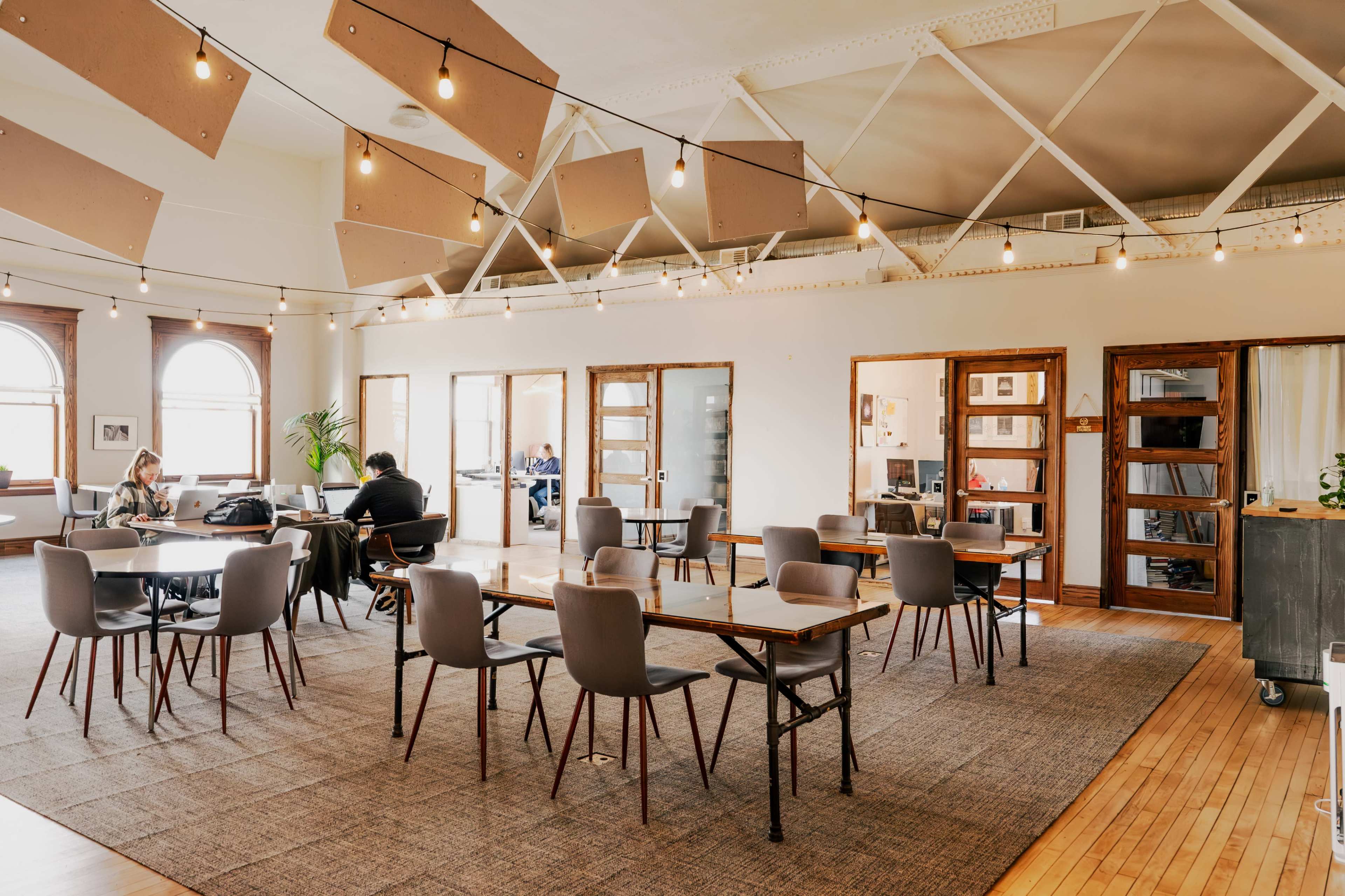 A spacious, well-lit coworking space with various tables and chairs, and people engaged in work activities.