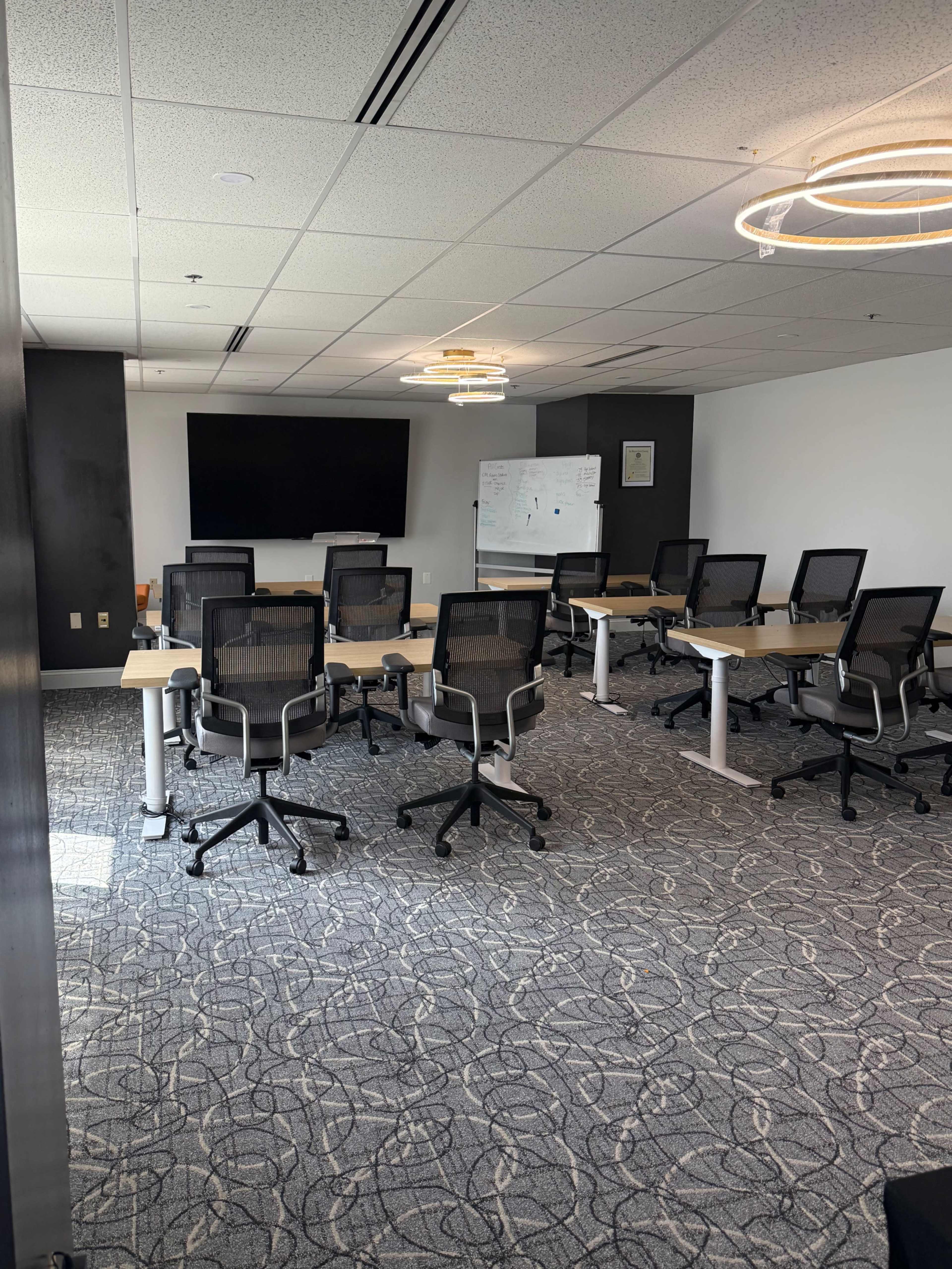 The image shows a modern conference room with several arranged chairs and tables, a large screen at the front, and a whiteboard on the side.
