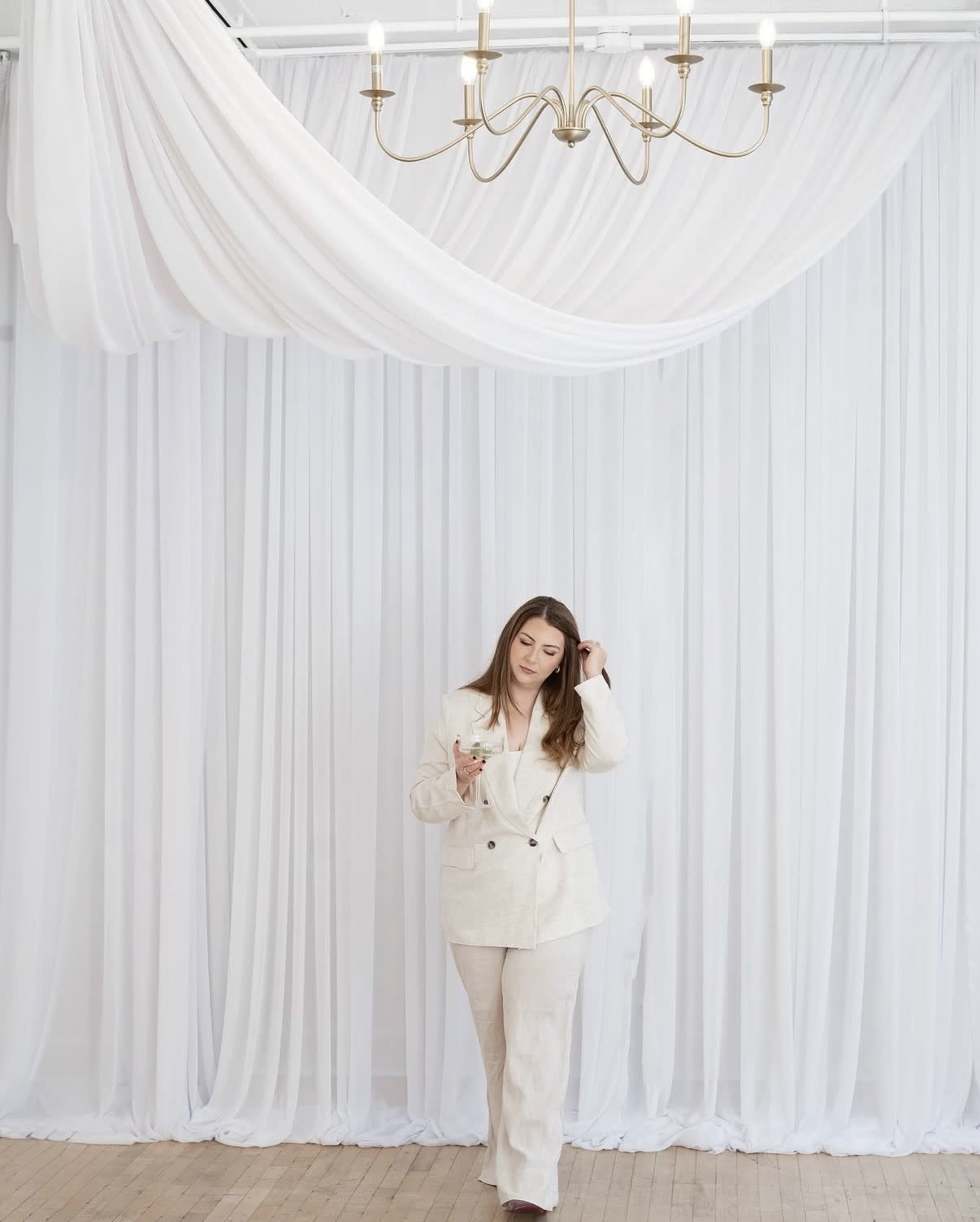 A woman in a white suit stands in front of a backdrop of sheer white drapery beneath a chandelier.