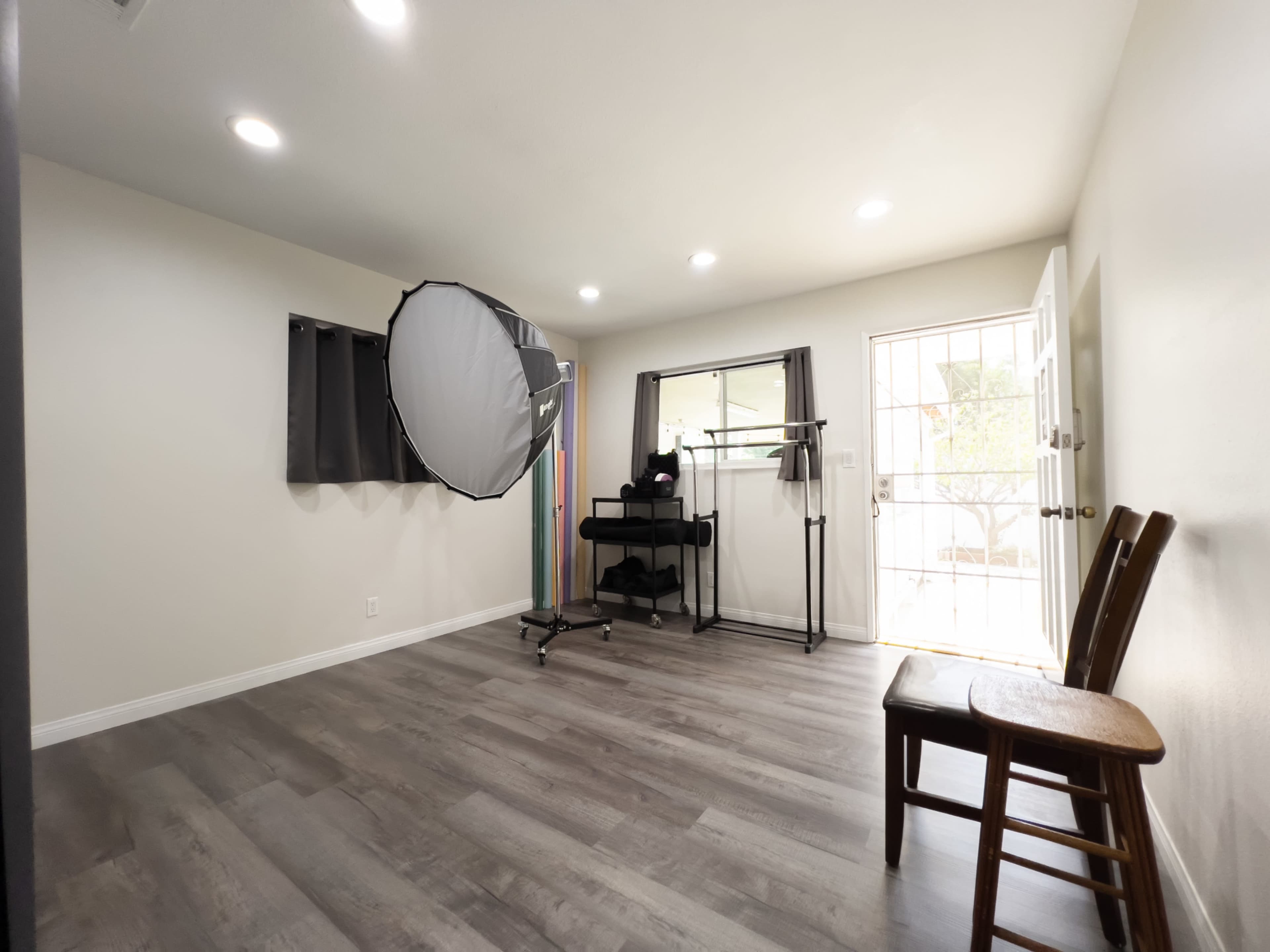 A room with light-colored walls, a wooden floor, a large circular softbox on a stand, a small table with a black cart, and a door leading to an outside area.