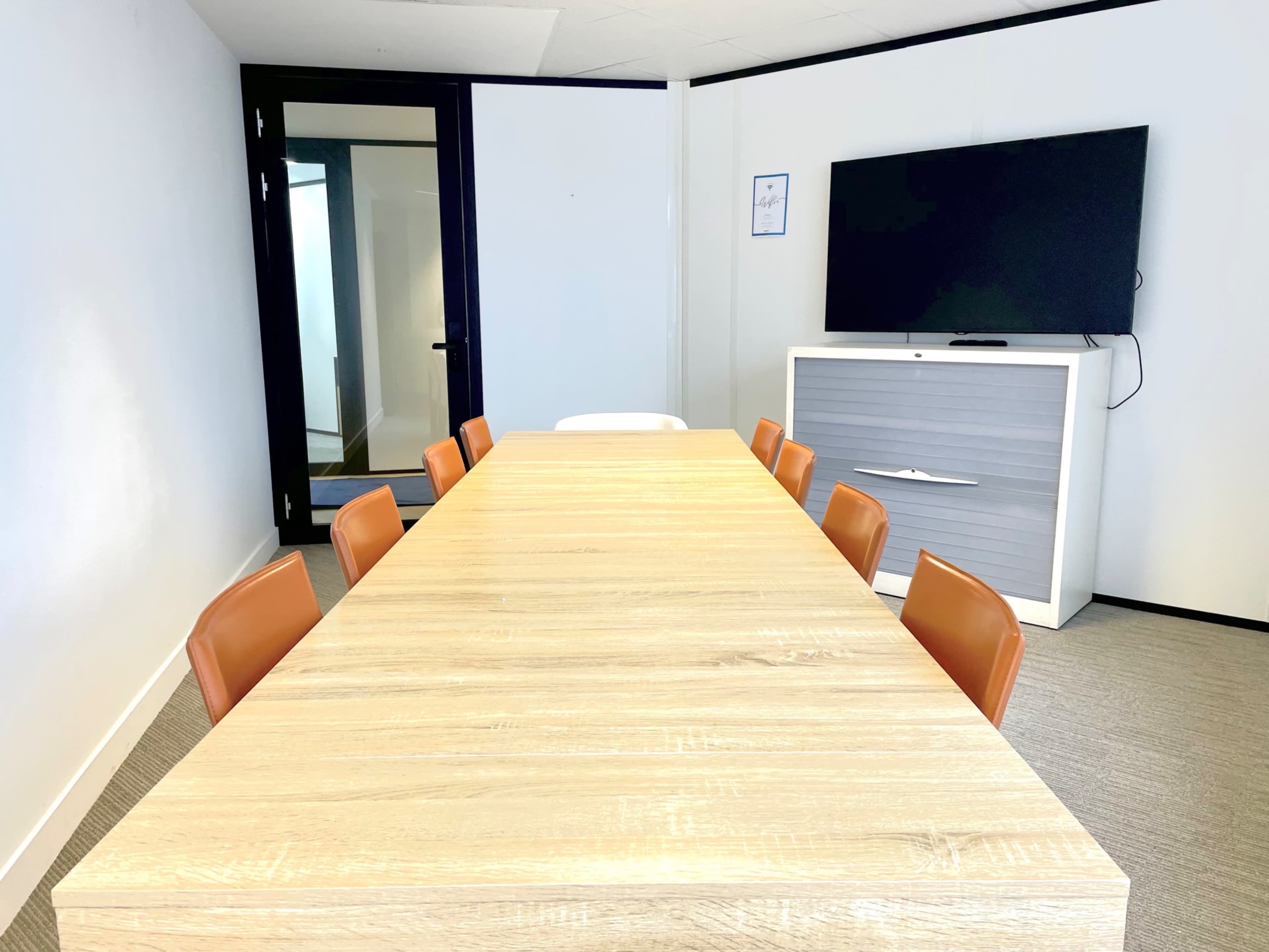 A long wooden conference table with eight orange chairs is positioned in a modern meeting room, facing a large wall-mounted television.