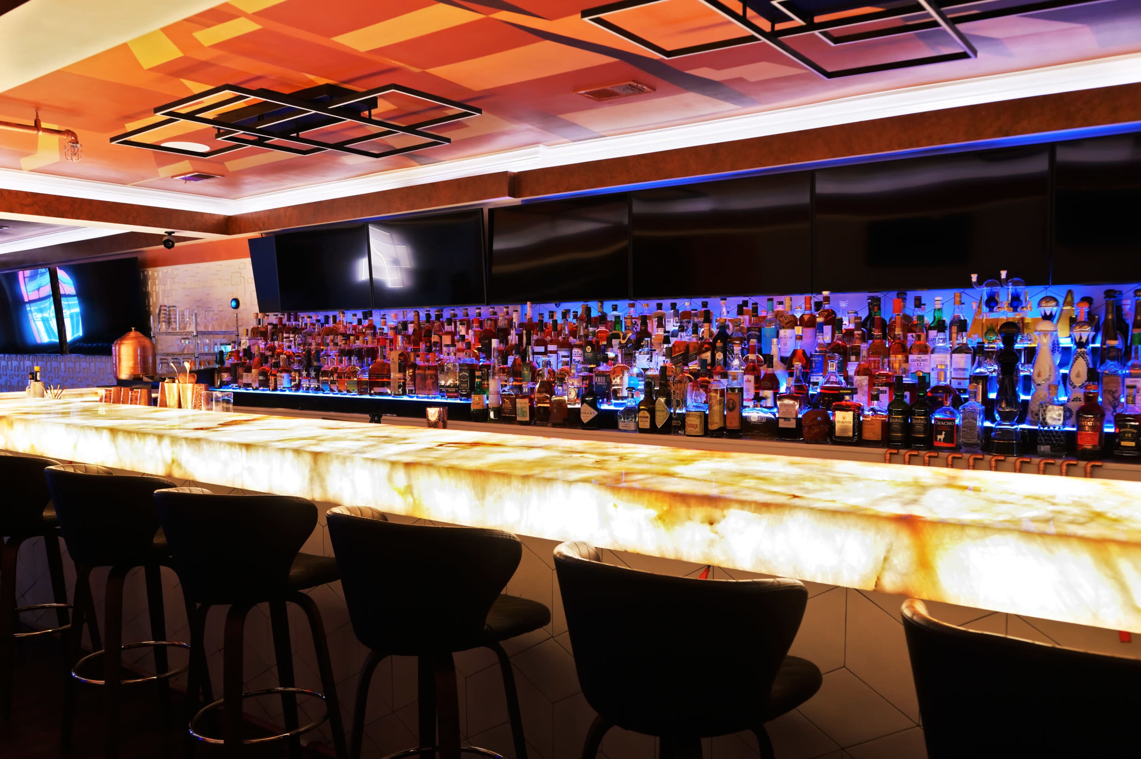 A sleek bar with a luminous onyx countertop, lined with high-backed stools, displays an array of illuminated bottles in the background.