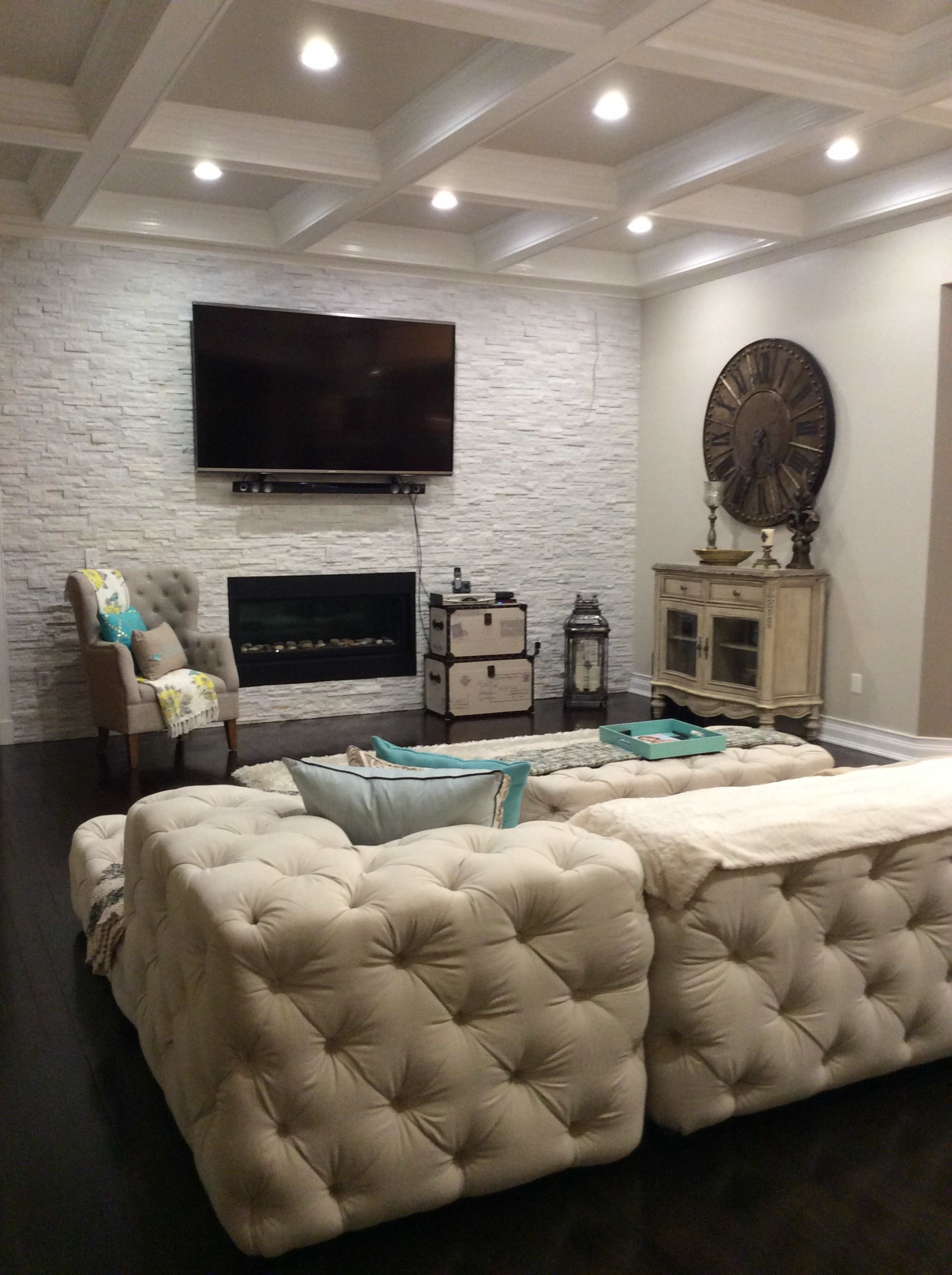 A modern living room featuring a tufted light-colored sofa, a wall-mounted television, a stone accent wall, and a decorative clock on a nearby cabinet.