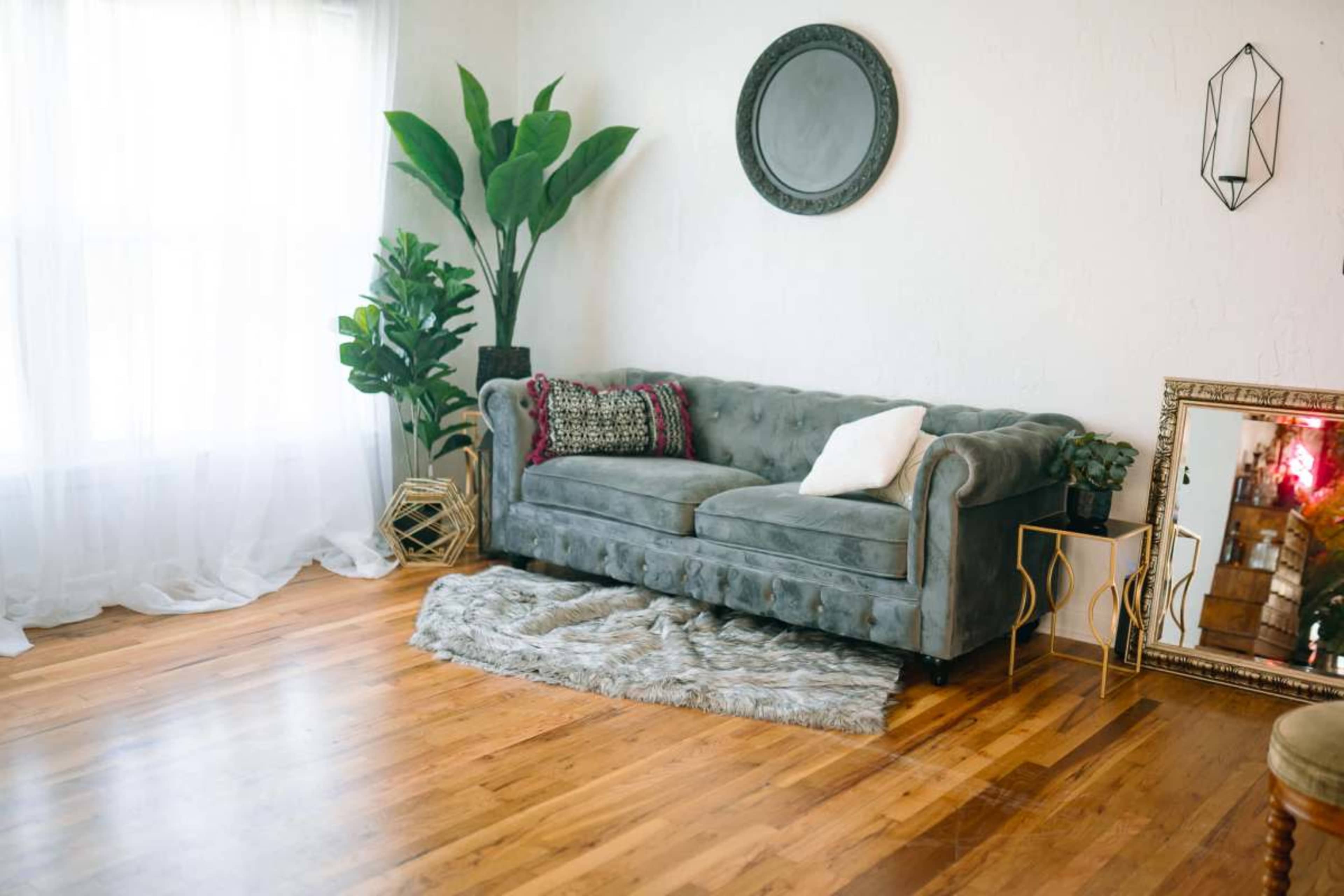 The image shows a spacious living room with a gray tufted sofa, a circular wall mirror, houseplants, and a decorative gold-framed mirror on the wooden floor.