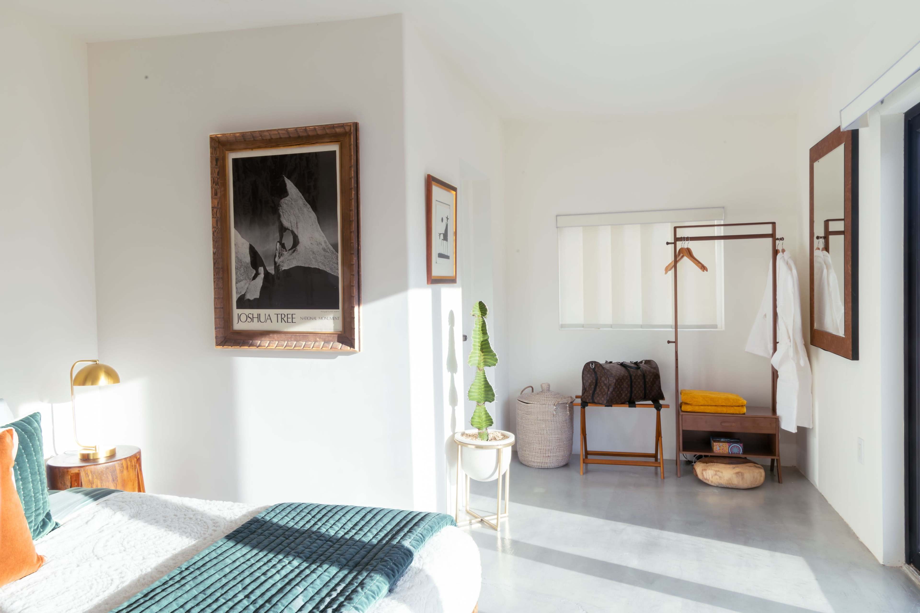 A bright and minimalist bedroom features a bed with a green throw, framed artwork on the wall, and a small seating area with a coat rack and woven basket.