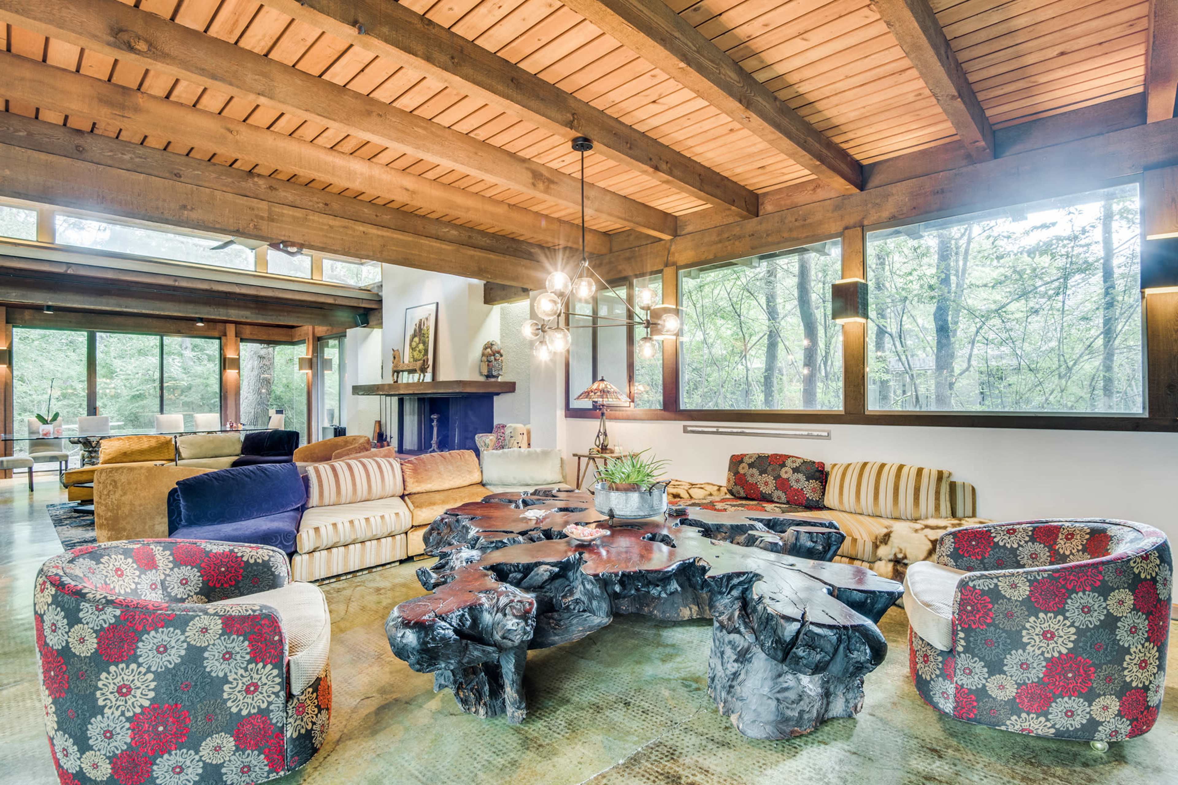 A spacious living room with large windows, showcasing a wooden beam ceiling and a unique coffee table made of dark, textured wood surrounded by patterned upholstered seating.