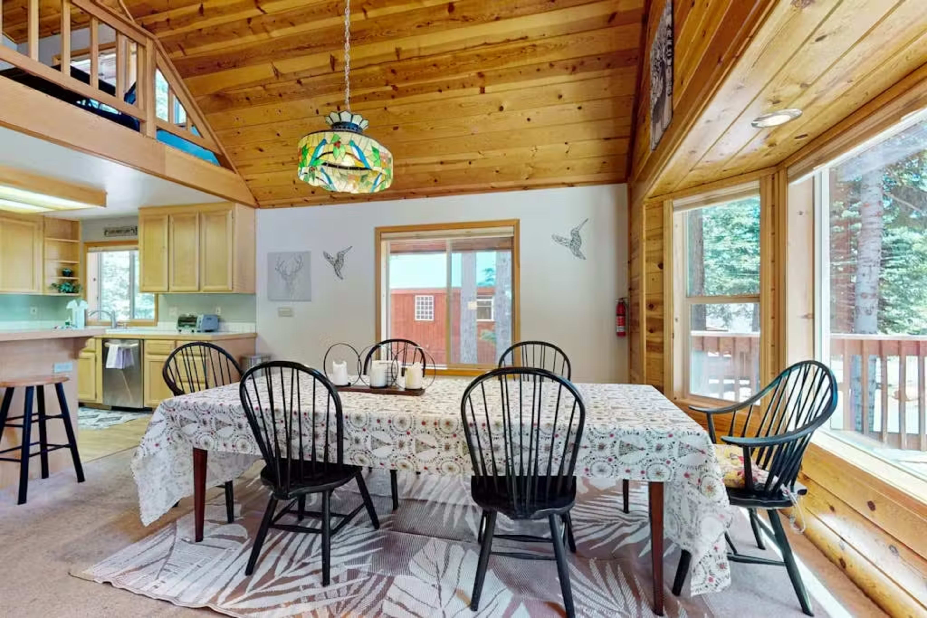 The image depicts a wooden dining area with a large table set for six, surrounded by black chairs and illuminated by a stained glass pendant light, with large windows offering views of the outdoors.