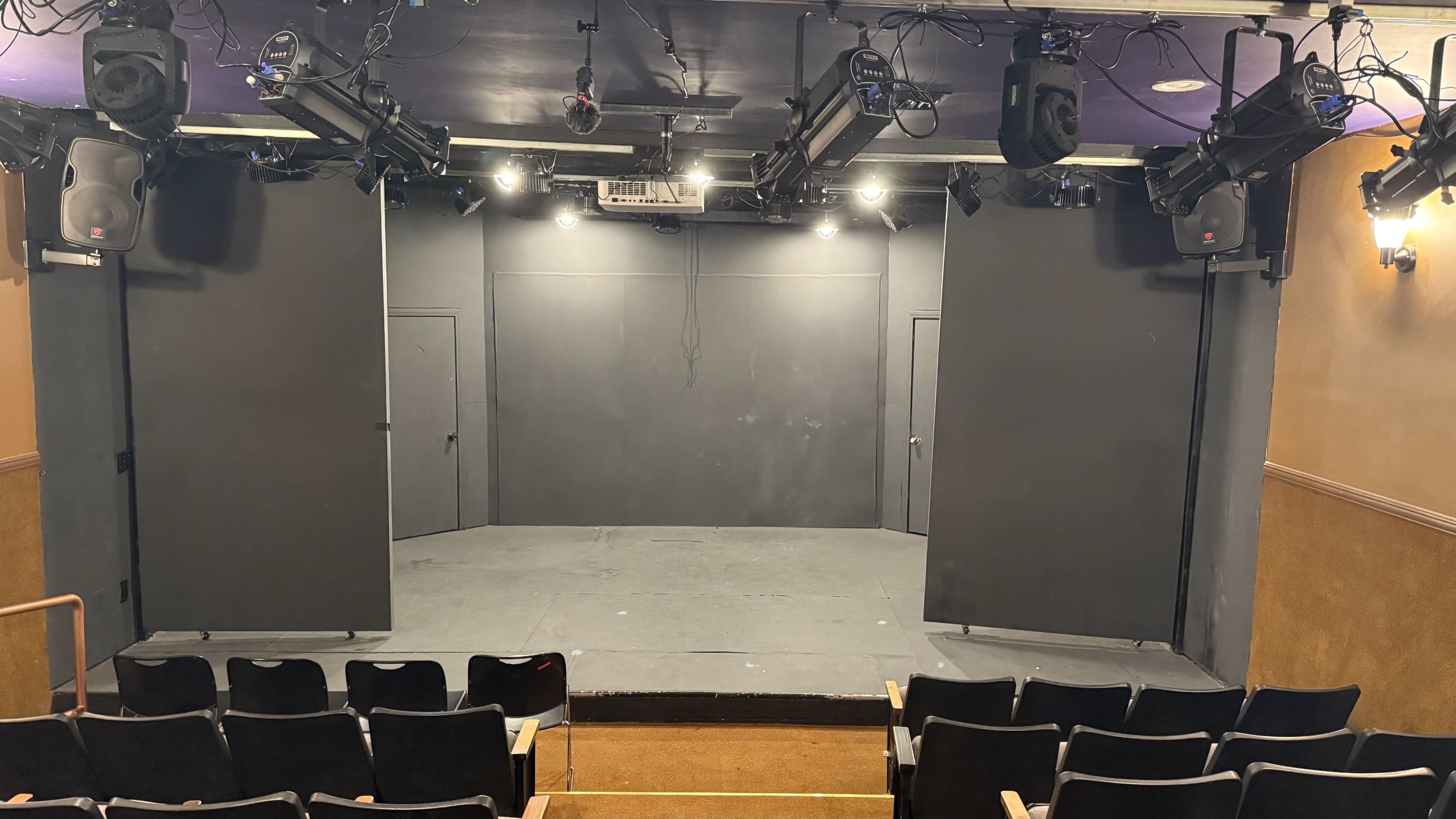 The image shows a small theater stage with black curtains, two side walls, and rows of empty seats facing the performance area.