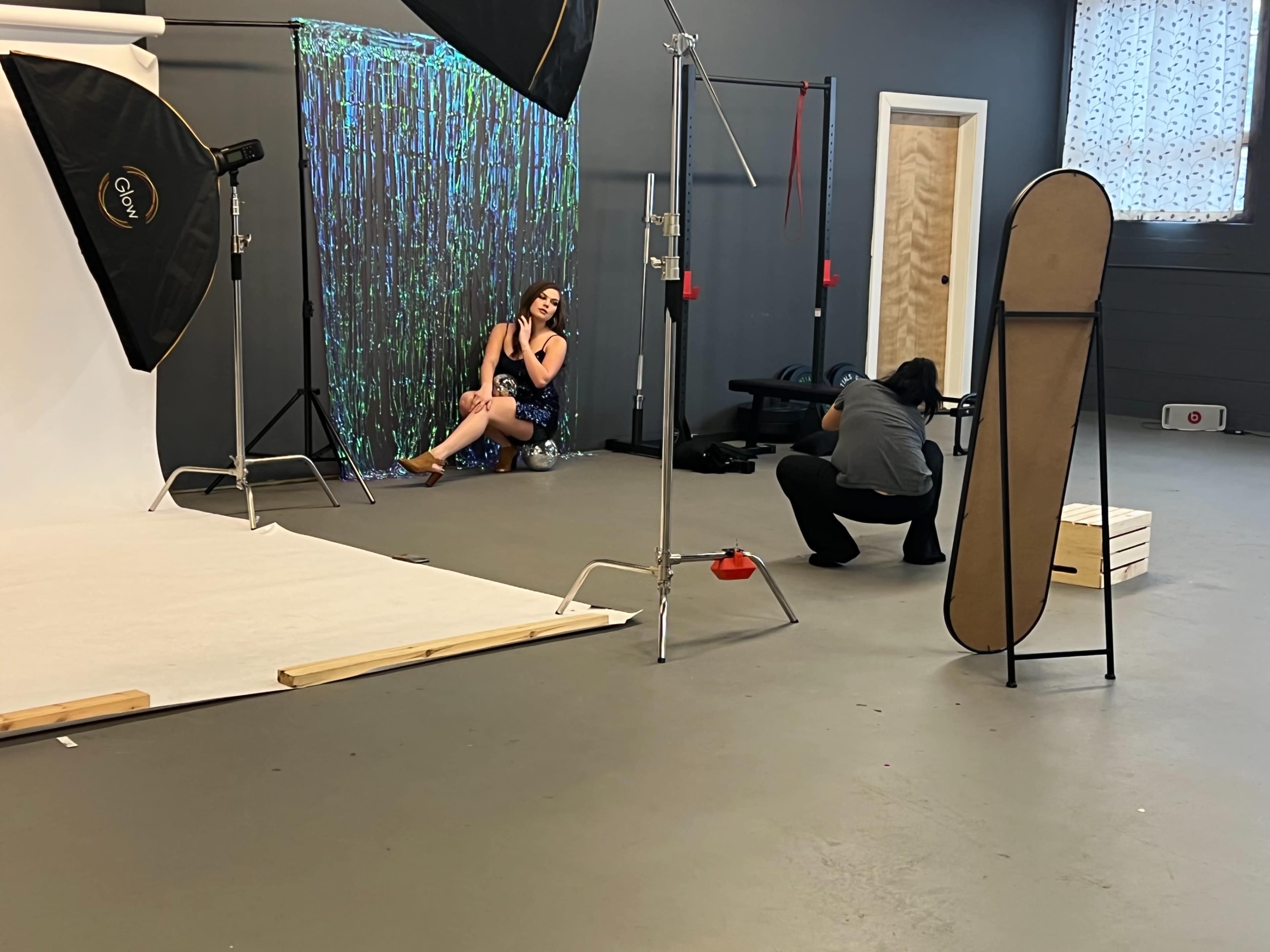 A photoshoot is taking place in a studio, featuring a model posed against a colorful backdrop while a photographer captures images.