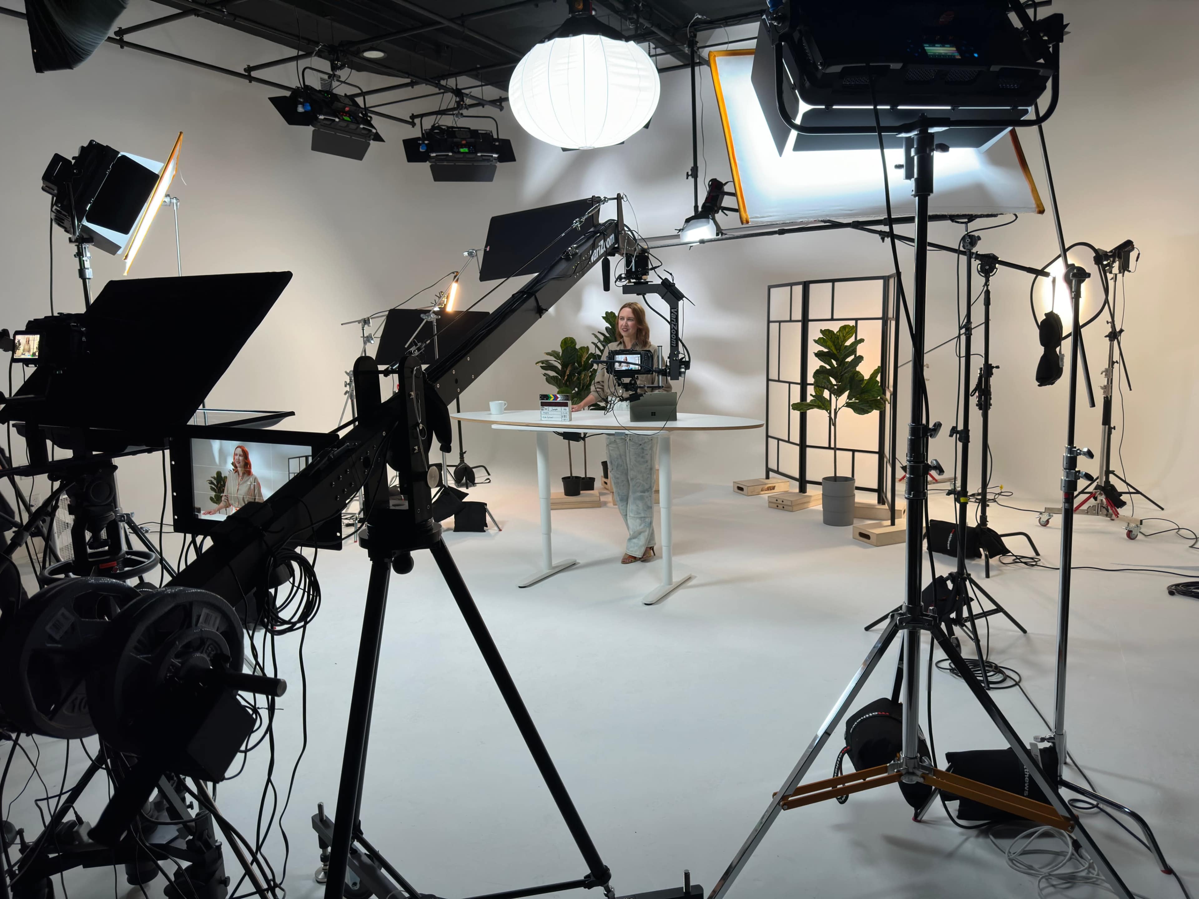A woman stands at a table surrounded by lighting equipment and potted plants in a studio setting.