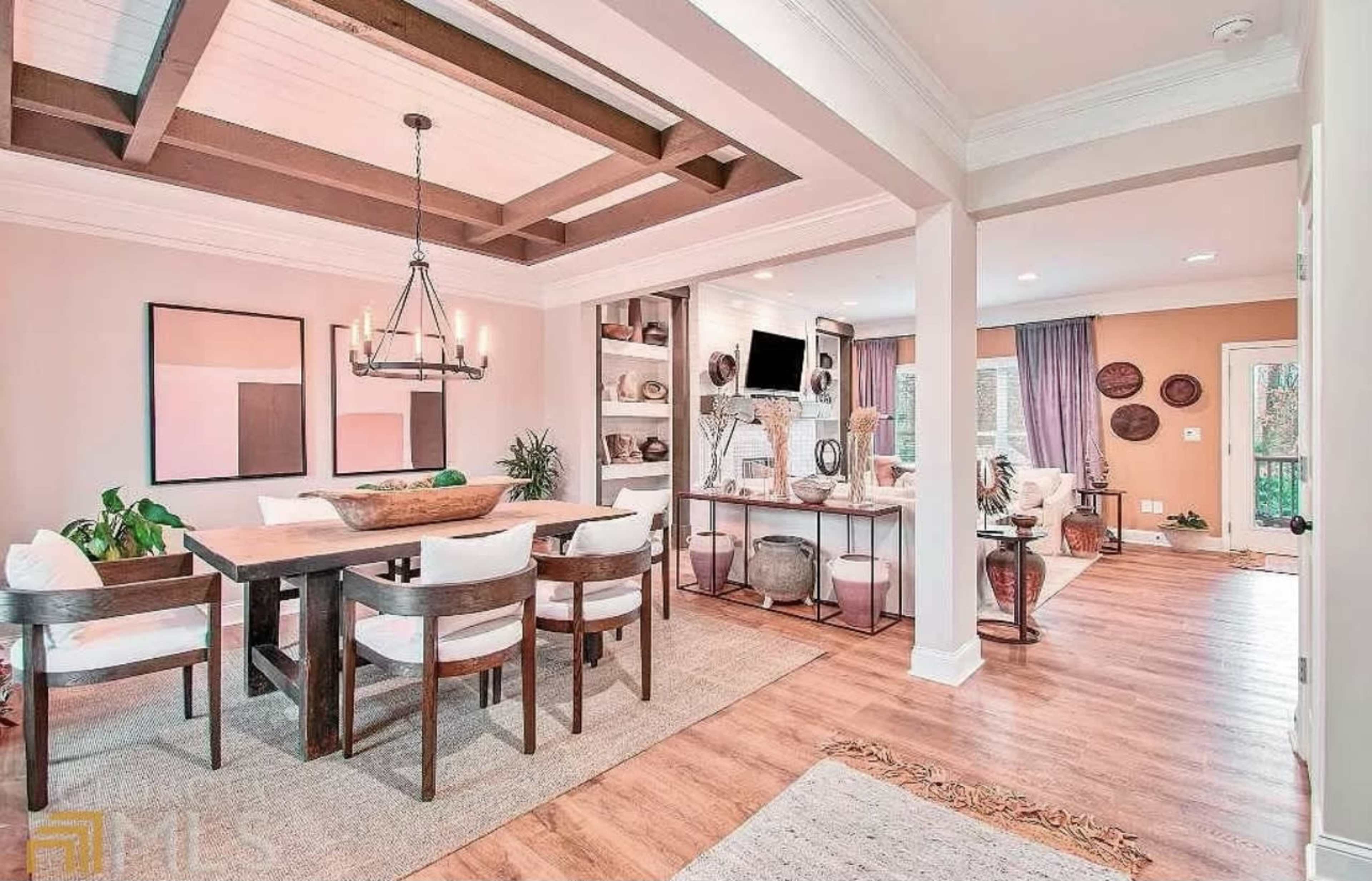 The image shows a dining area with a wooden table and white chairs, adjacent to a living room featuring a television and shelves.
