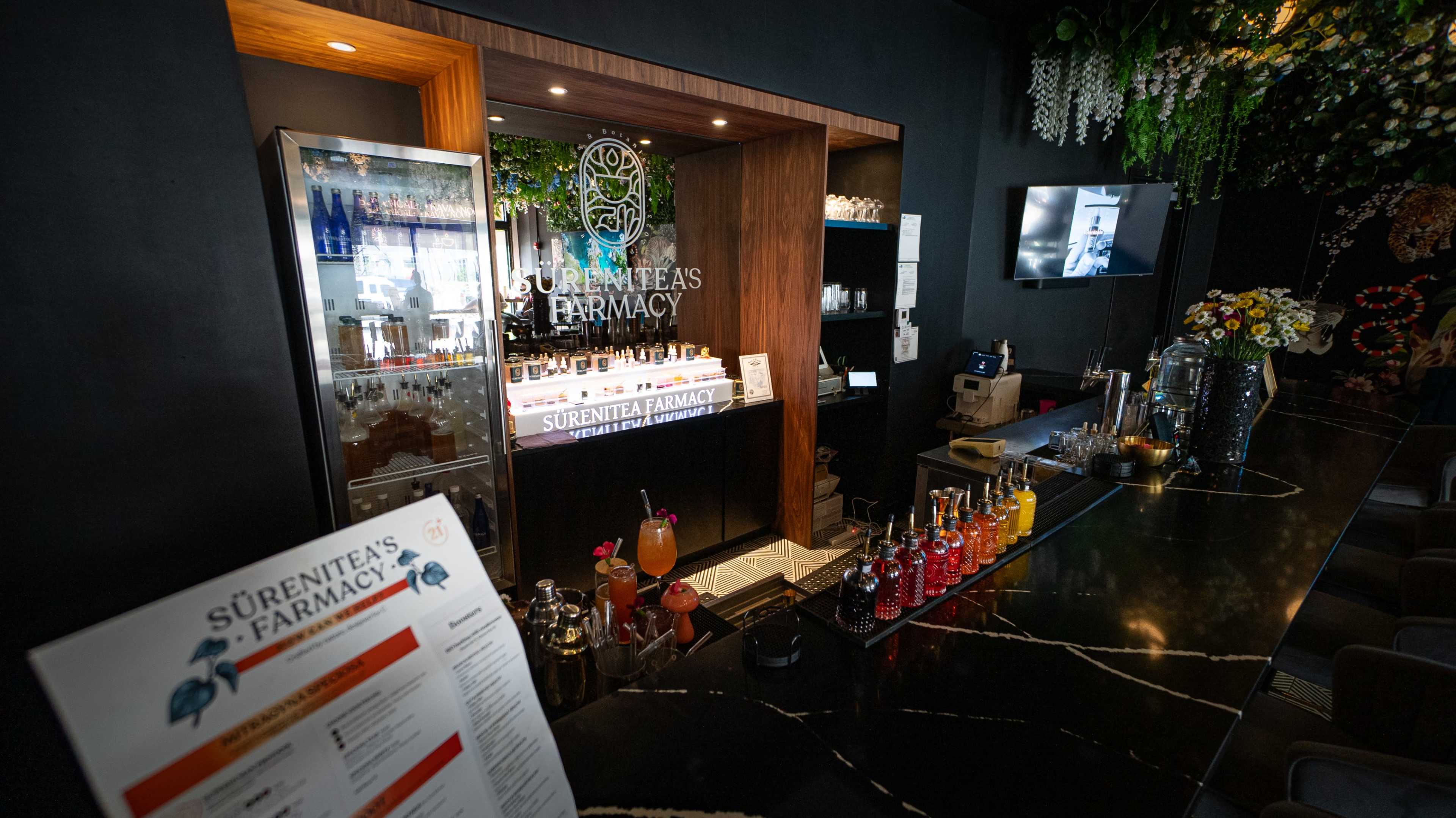 The image shows a modern pharmacy bar with a sleek counter, showcasing various colorful bottles and drinks, alongside a menu on the foreground.