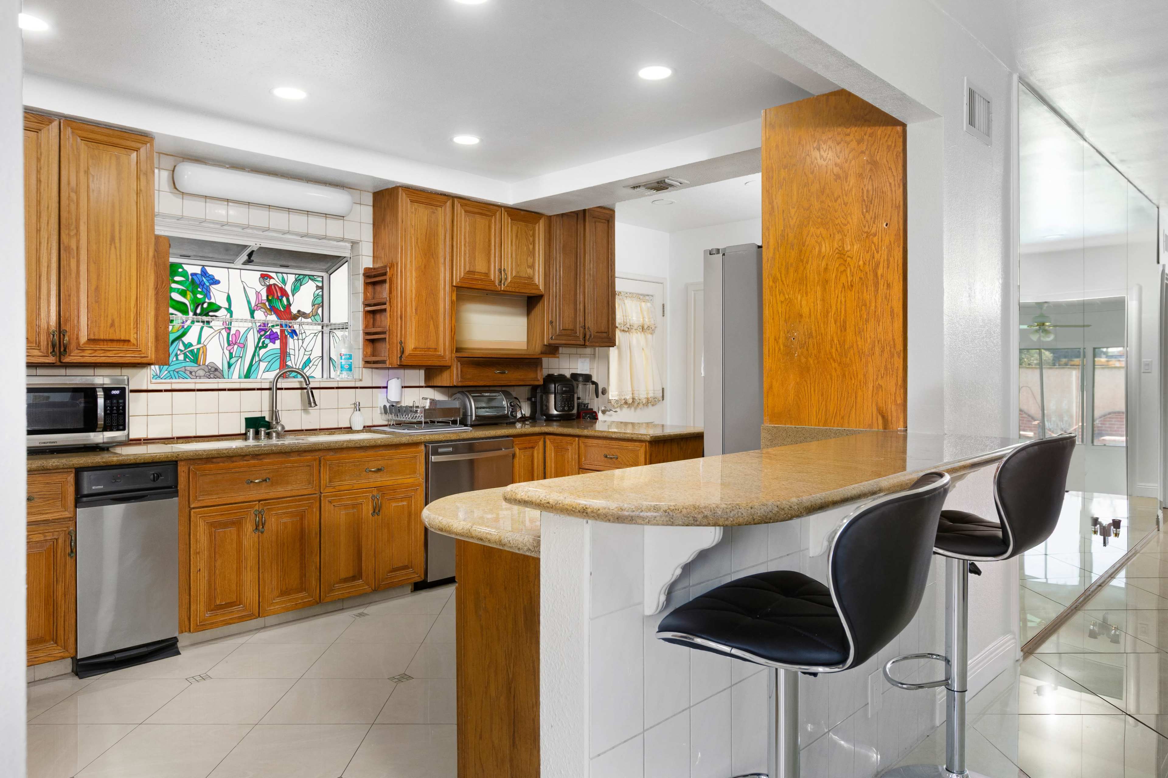 The image shows a spacious kitchen with wooden cabinets, stainless steel appliances, and a breakfast bar with black stools.