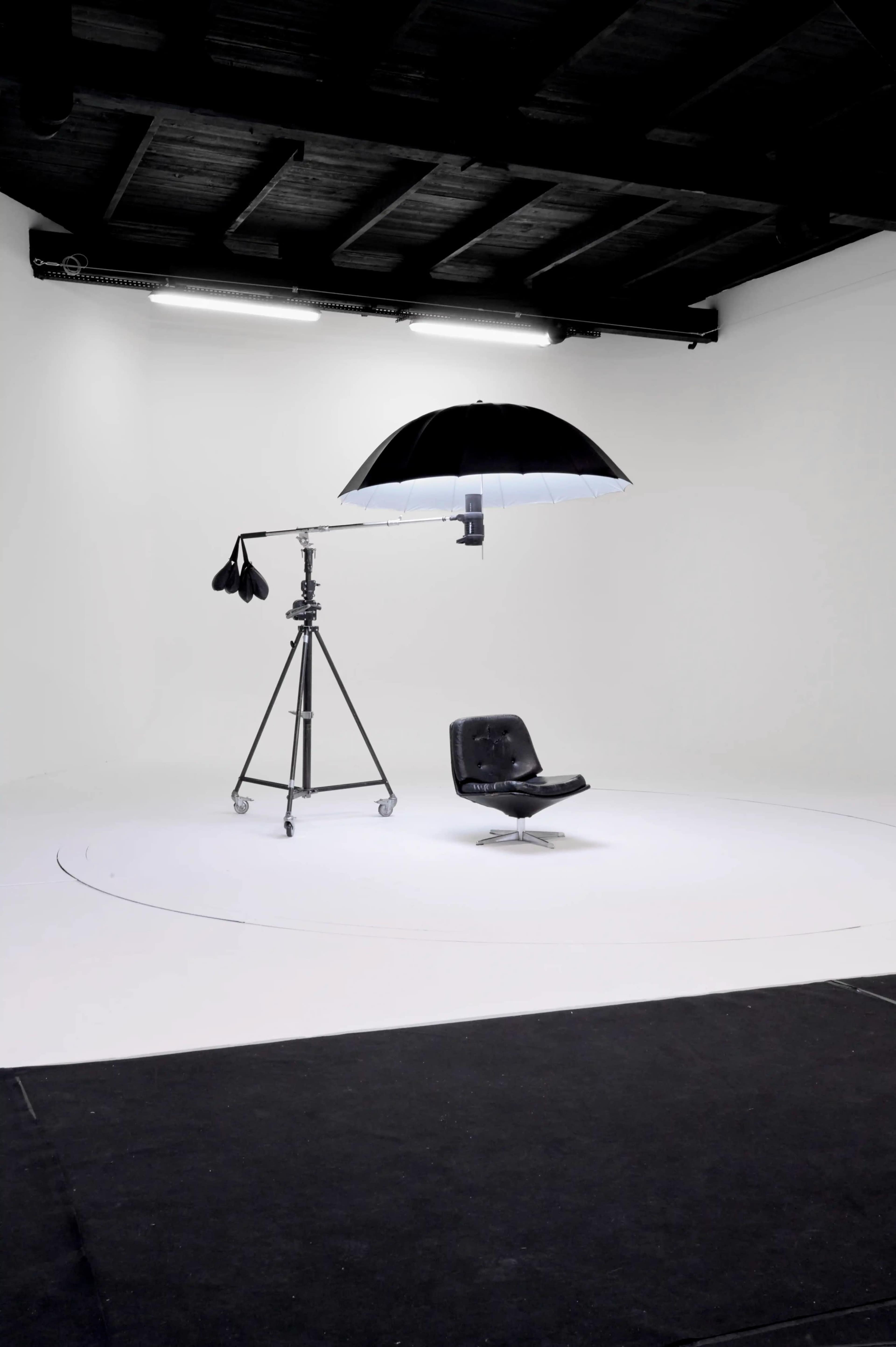 The image shows a minimalist photo studio setup with a black chair beneath a large umbrella light on a tripod.