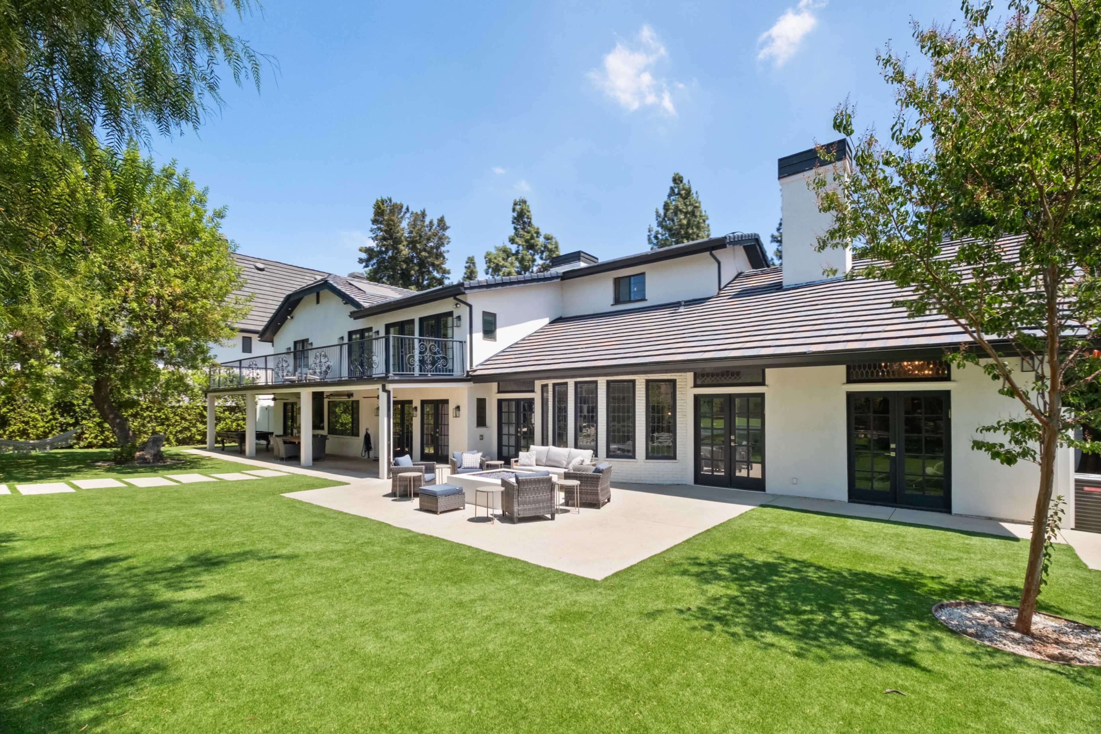 The image shows a modern two-story house with a spacious backyard featuring a patio area and landscaping under a clear blue sky.