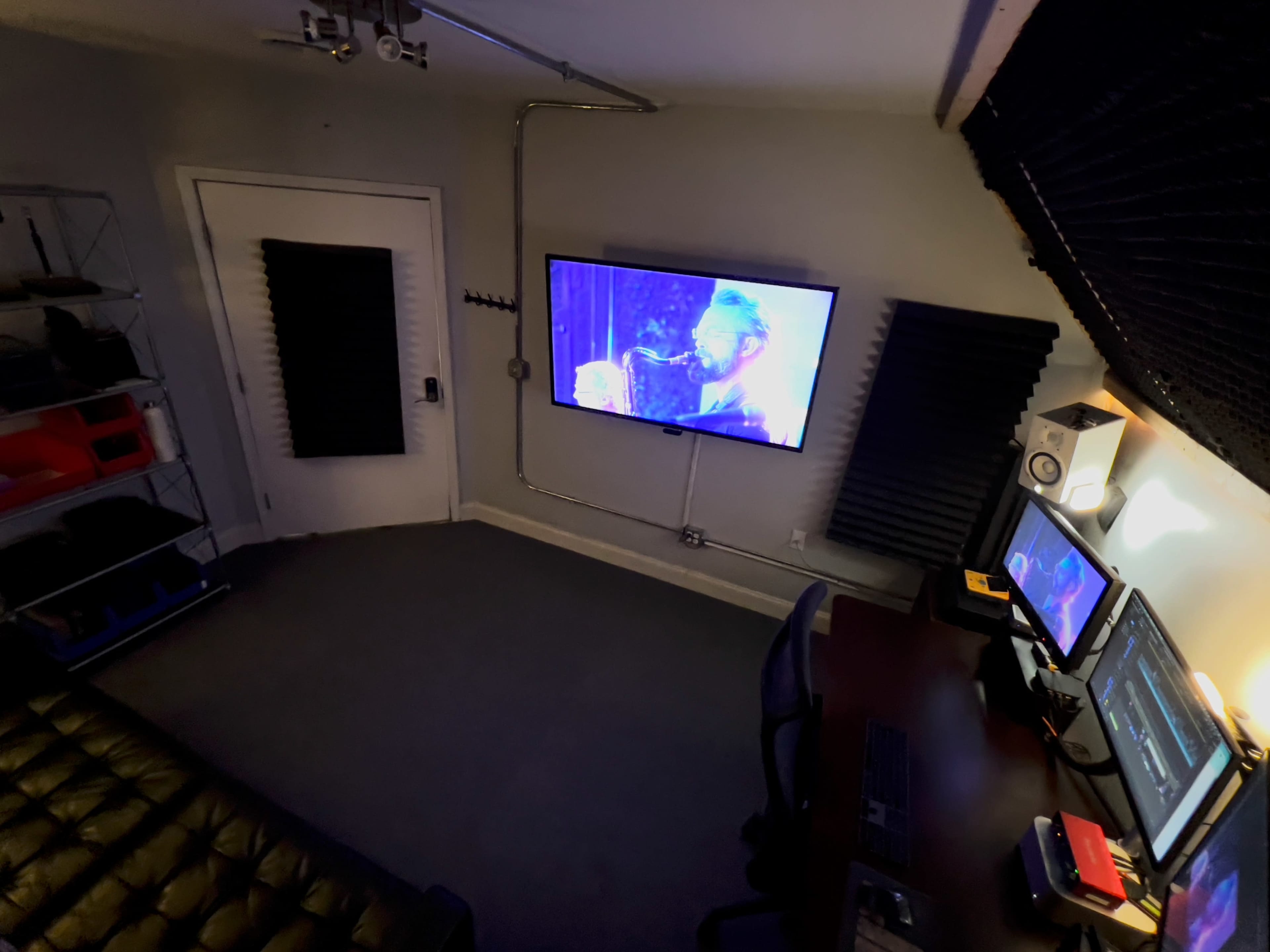 A dimly lit room features a large screen displaying a video, a desk with computers, and soundproofing panels on the walls.
