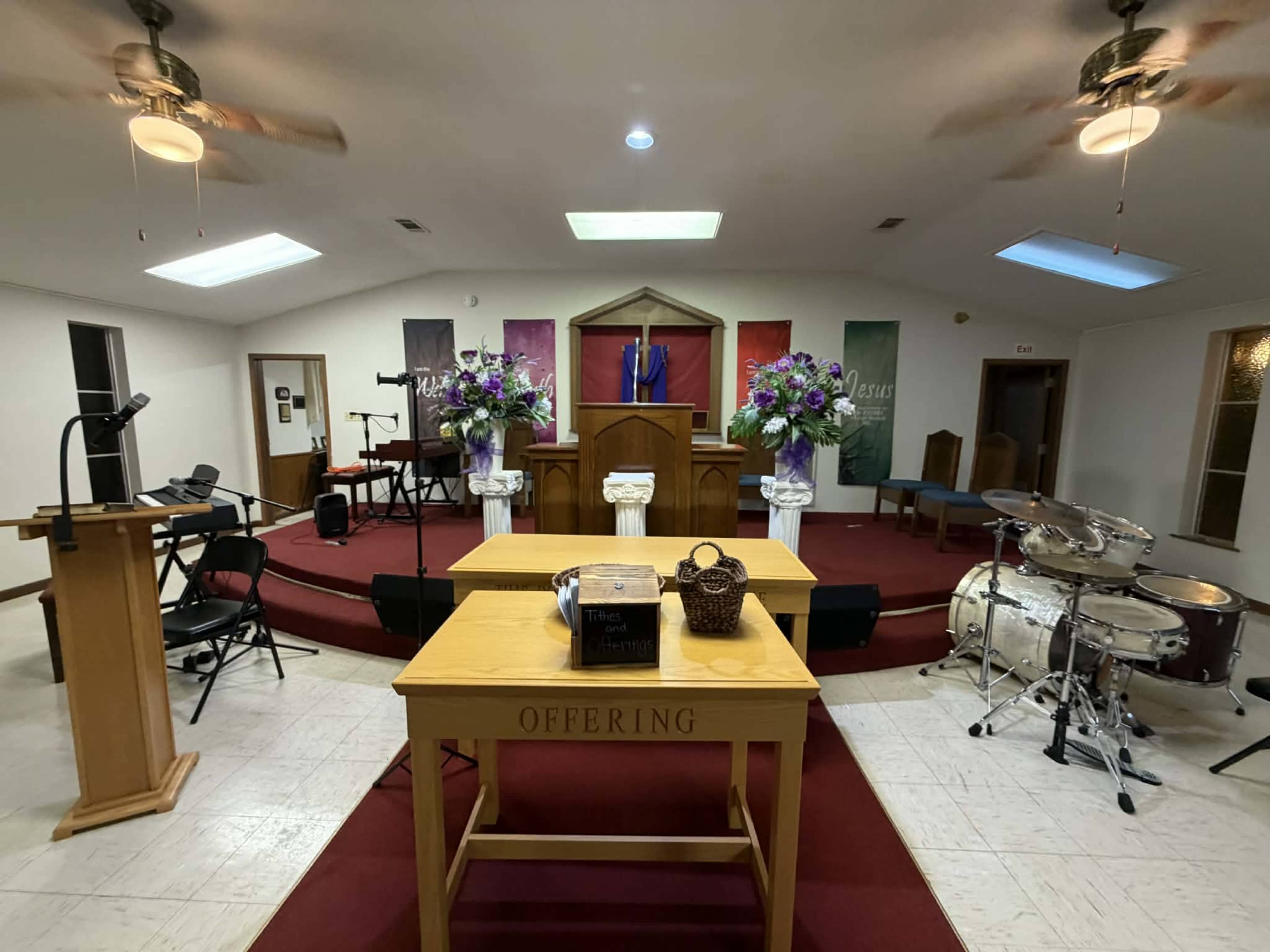 The image shows a church interior featuring a wooden offering table with flowers, a pulpit in the background, and musical instruments to the side.
