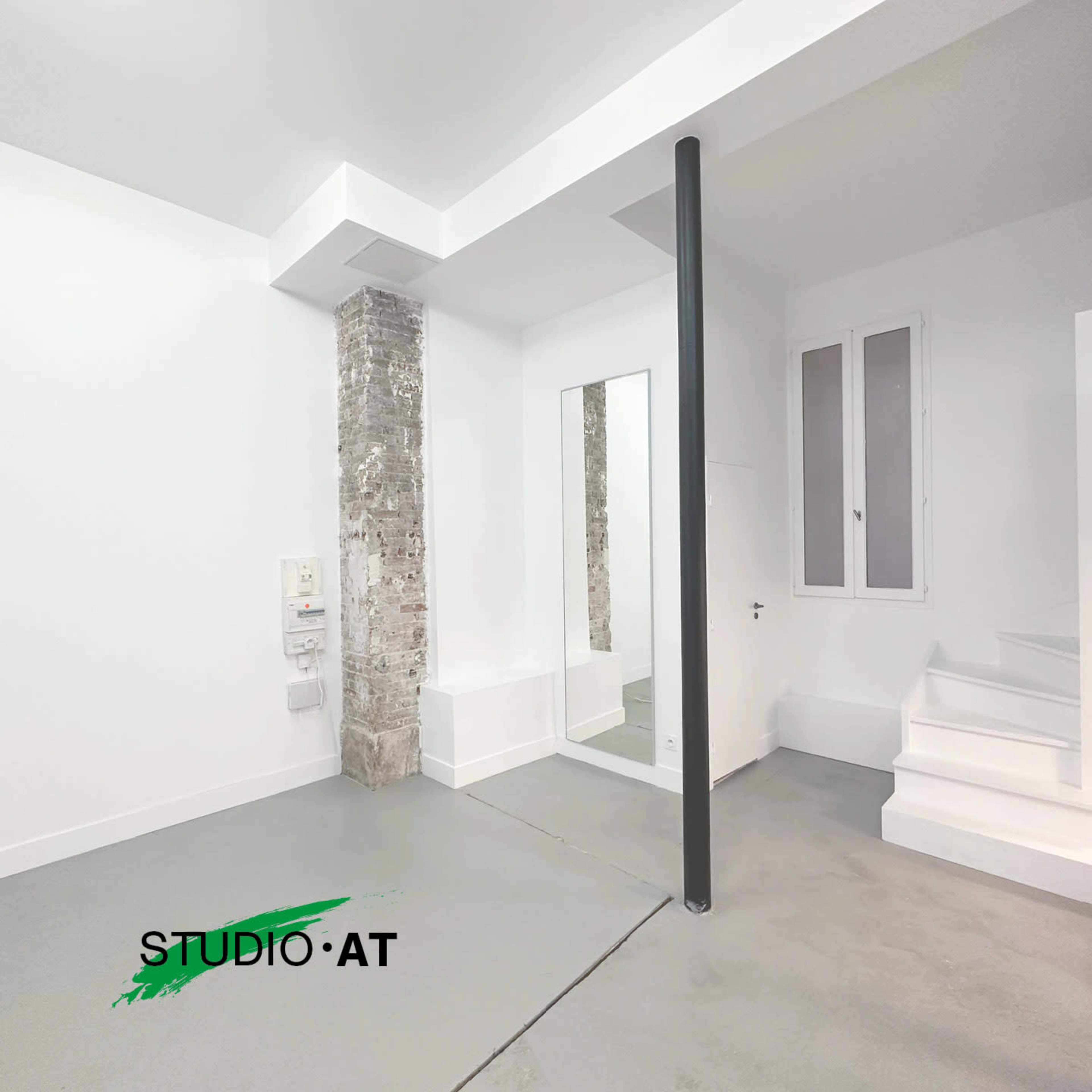 The image shows a minimalist interior space with a light gray floor, white walls, a full-length mirror, and a column along with a staircase in the background.