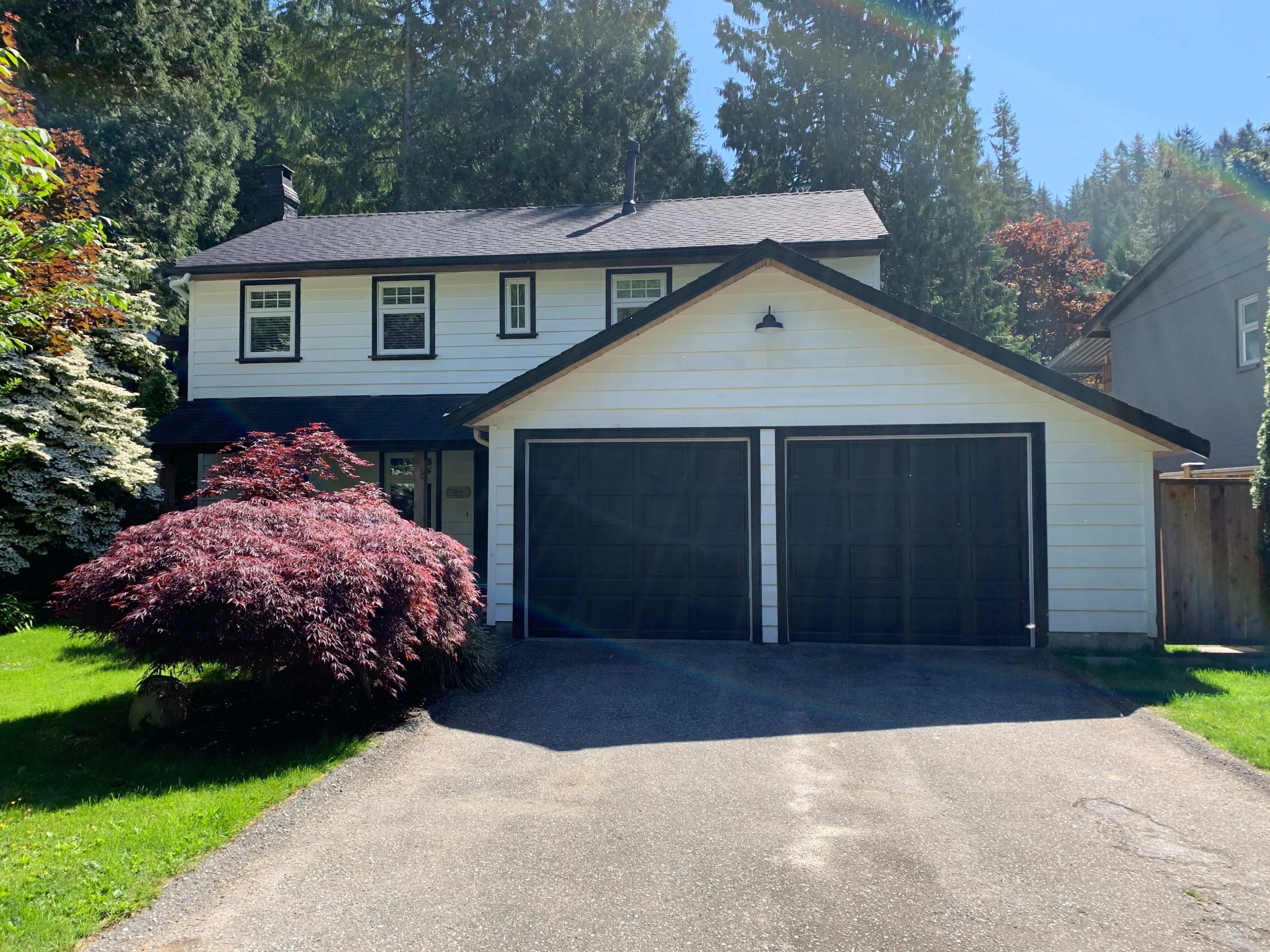 The image shows a two-story house with a black roof, white siding, and a double garage, surrounded by tall trees and shrubs.