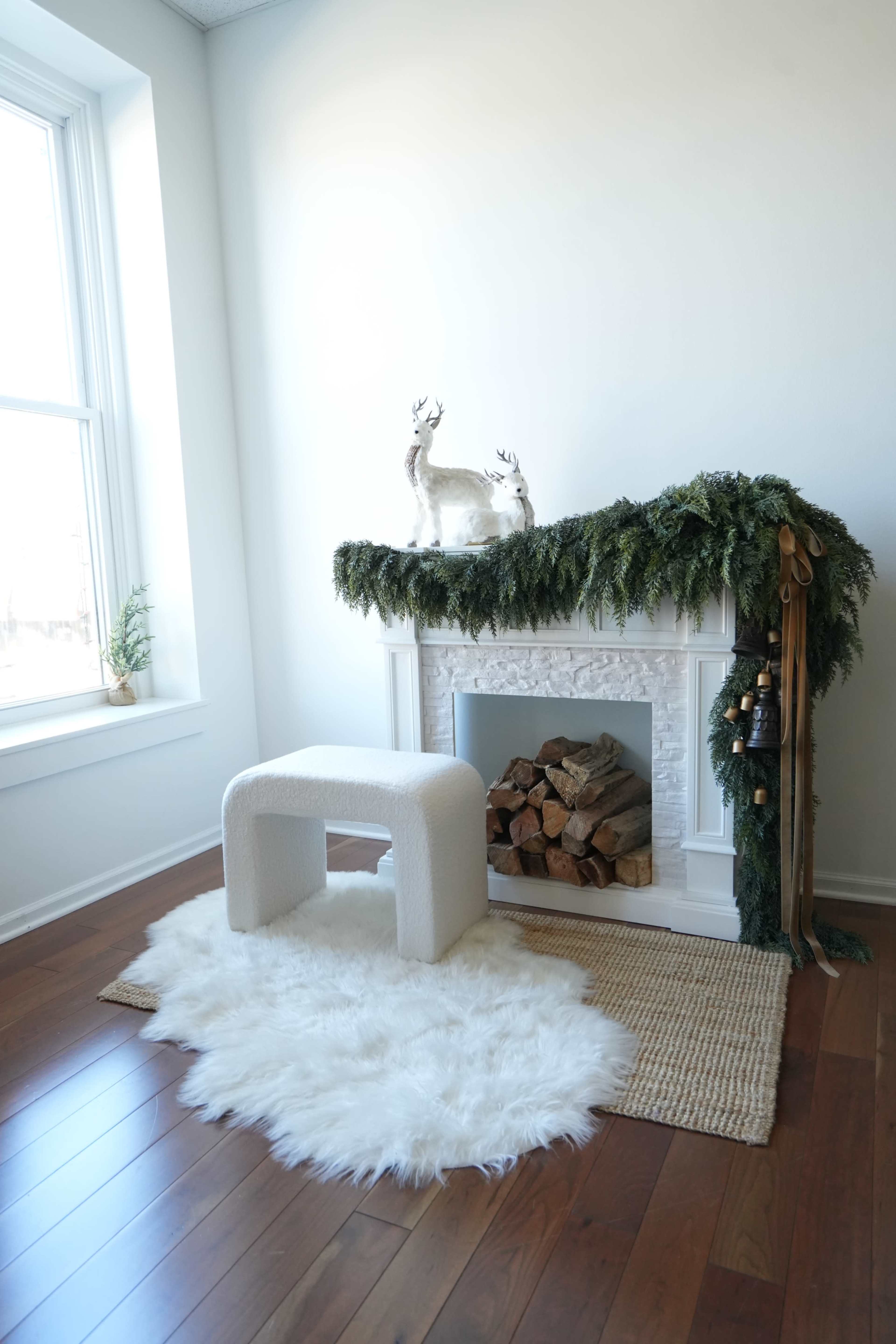 A minimalist fireplace is decorated with greenery, two deer figurines, and a stack of firewood, accompanied by a white rug and a small, soft stool.