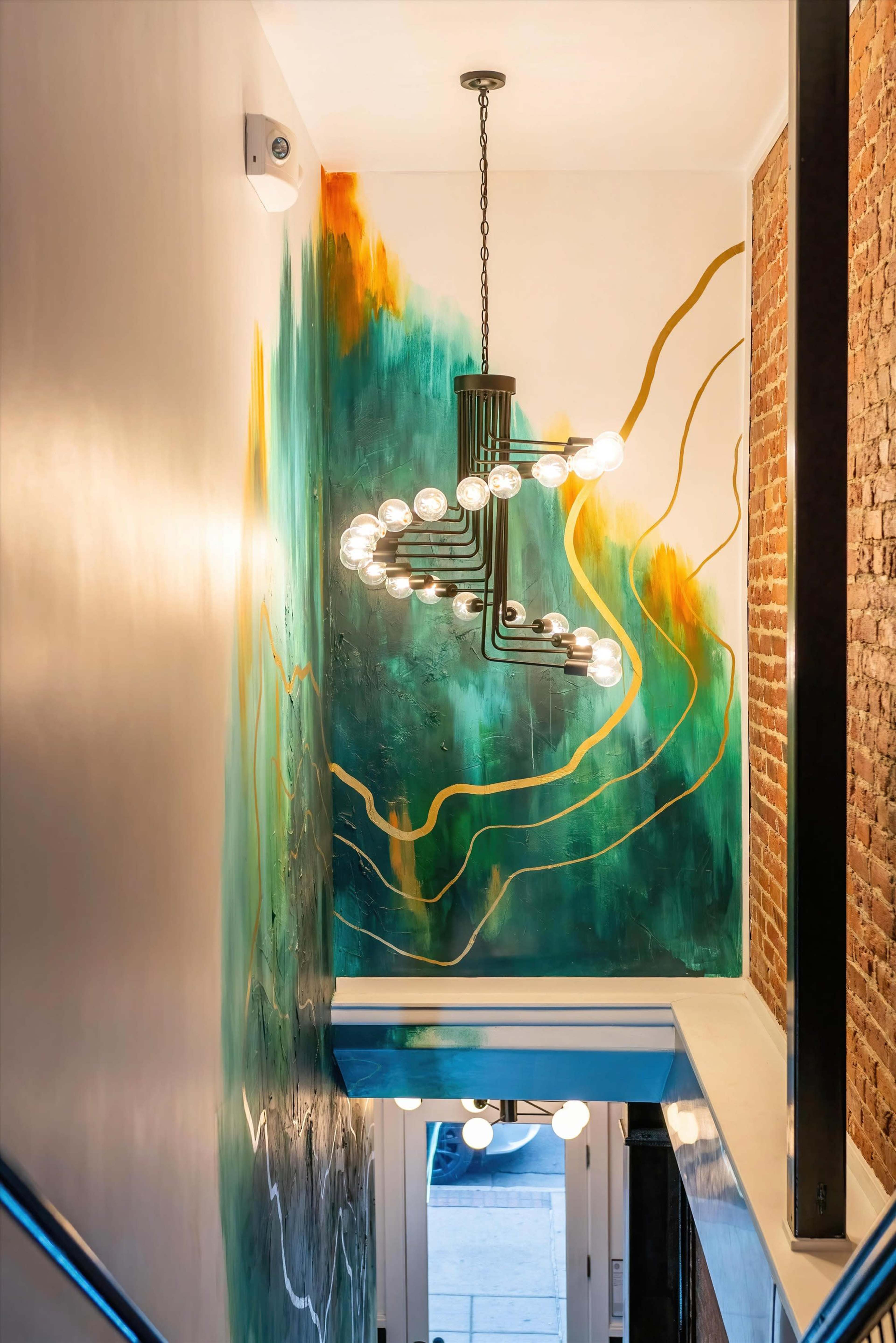 A colorful mural with green and gold accents adorns the wall of a well-lit stairway featuring a modern chandelier.