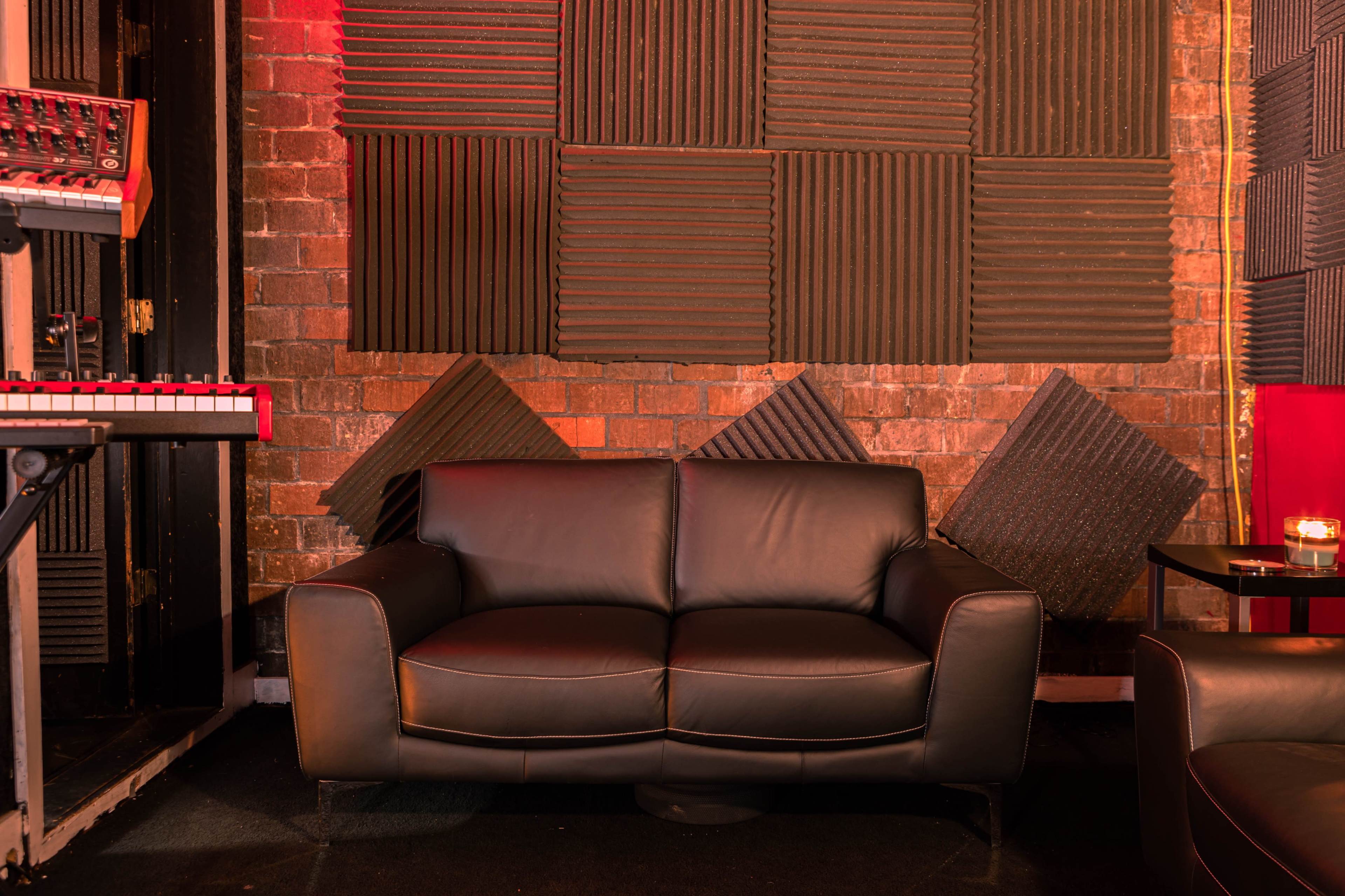 A leather couch is positioned against a brick wall with acoustic panels and musical equipment nearby.