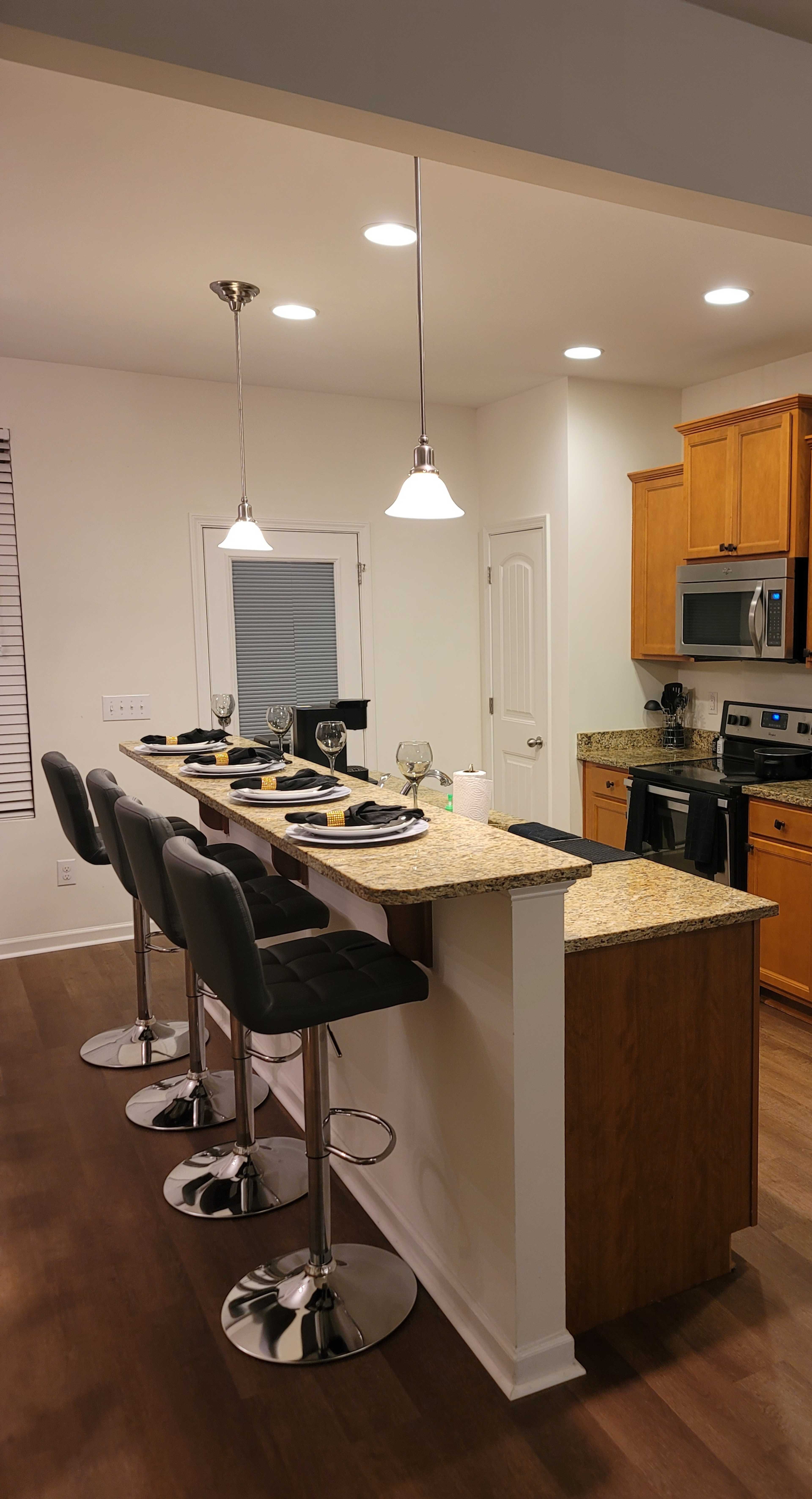 A modern kitchen features a bar area with four high stools, granite countertops, and pendant lights above, adjacent to the cooking area with wooden cabinets and stainless steel appliances.