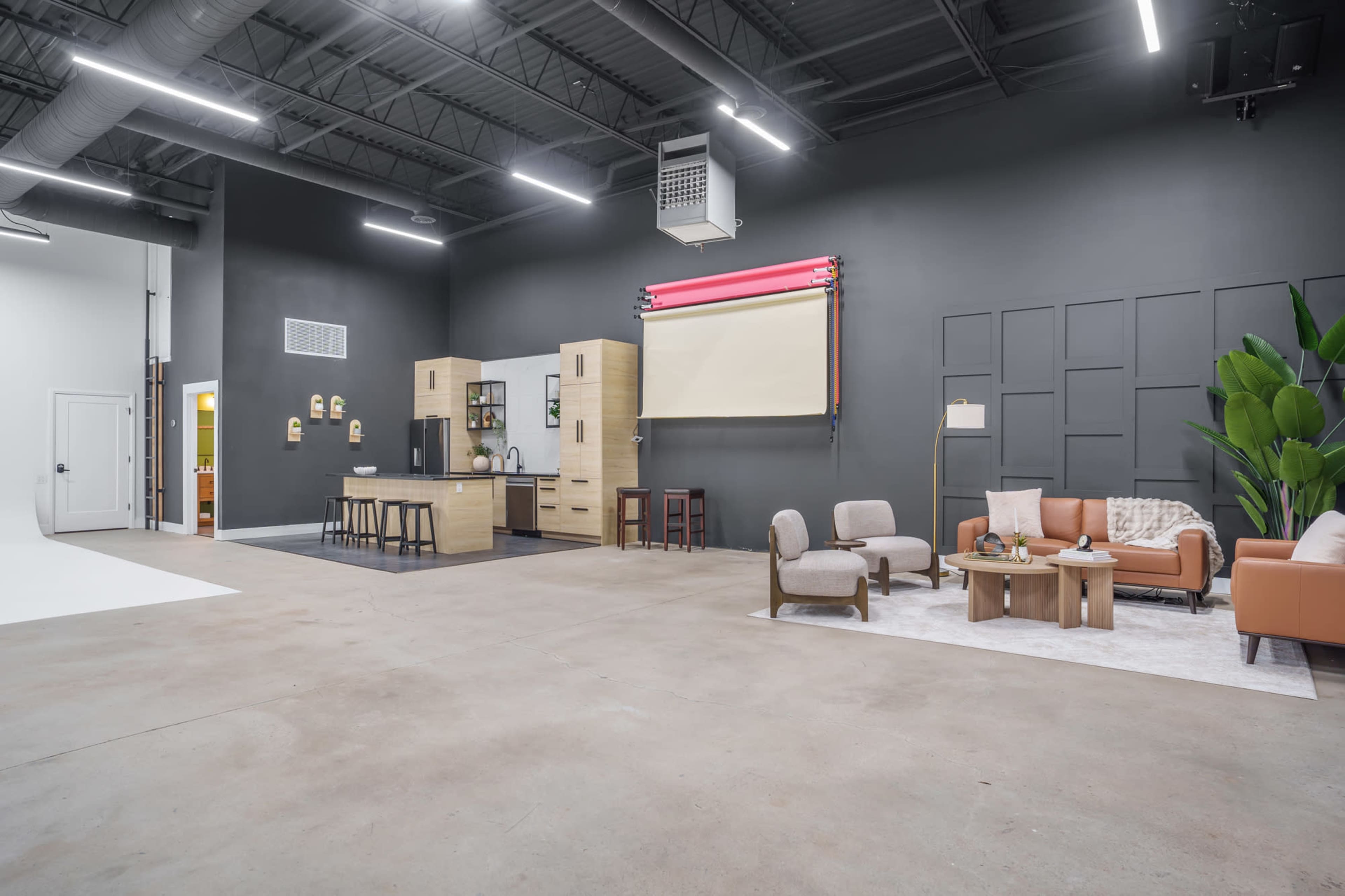 The image shows a spacious, modern studio featuring a kitchen setup, seating area, and a large dark wall with decorative elements.