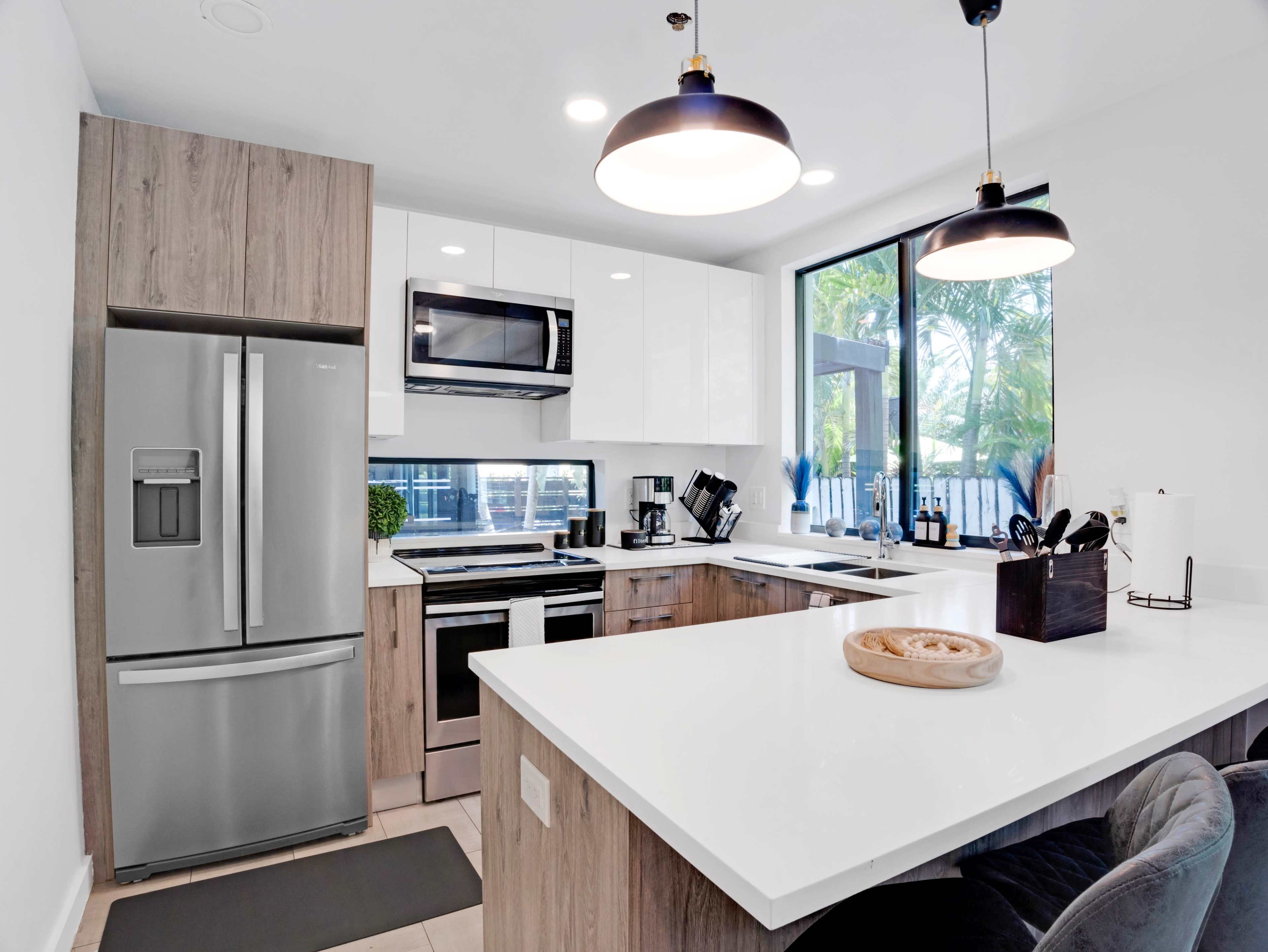 The modern kitchen features stainless steel appliances, a large island with a wooden serving platter, and a window overlooking greenery.