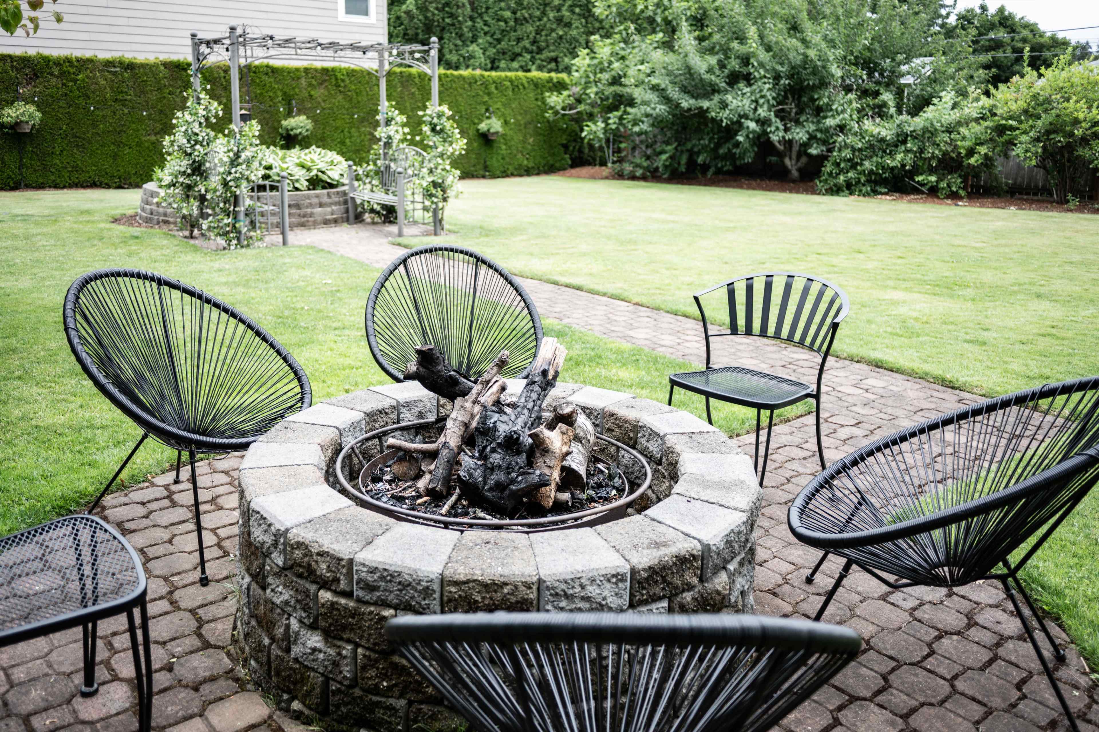 A circular stone fire pit surrounded by black wire chairs in a grassy backyard with a landscaped garden feature in the background.