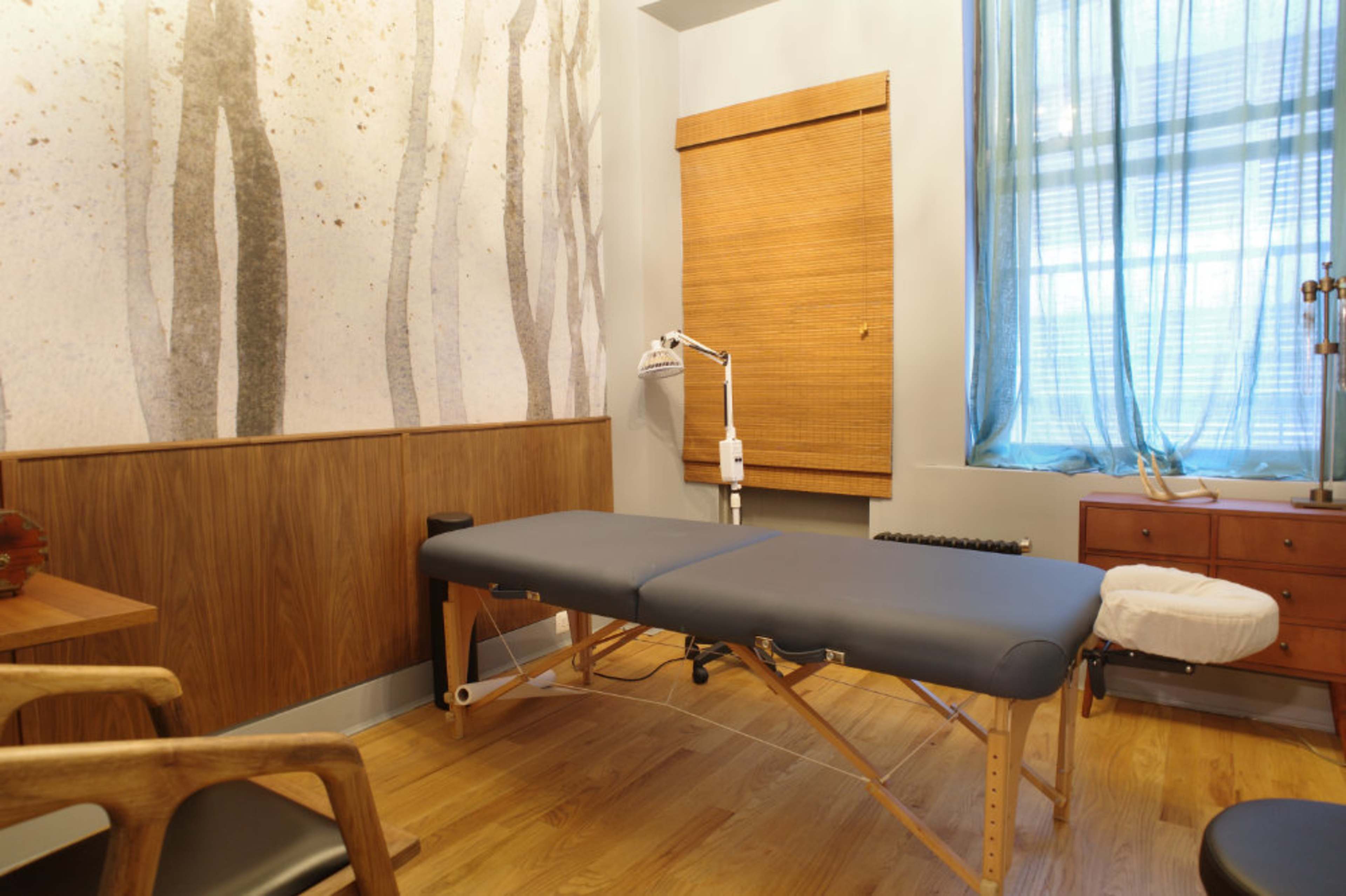 A massage therapy room features a wooden table, a chair, and a large window with sheer curtains.