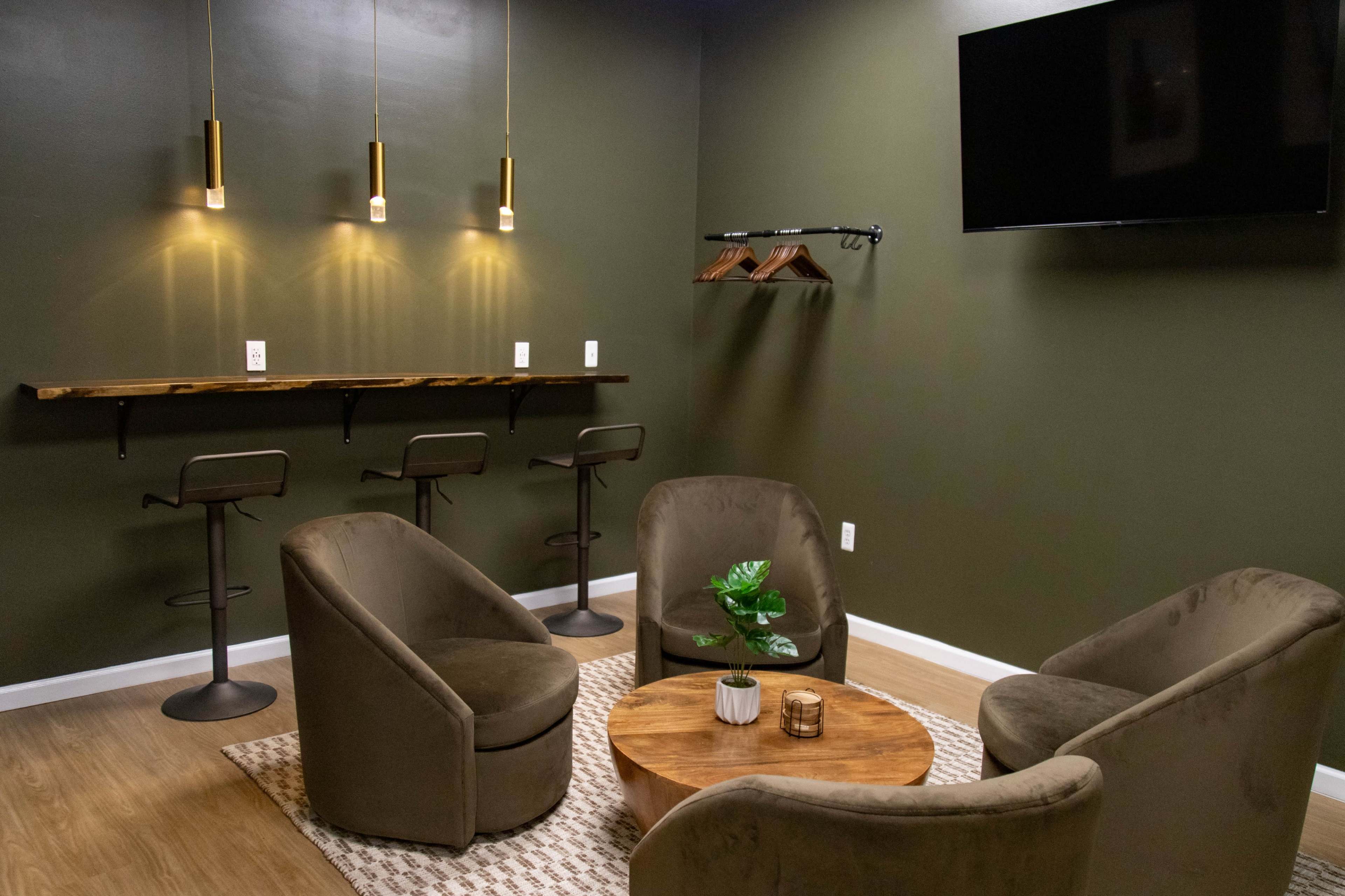 The image shows a modern lounge area with four round armchairs arranged around a wooden coffee table, a wall-mounted TV, and a bar with two high stools against a dark green wall.