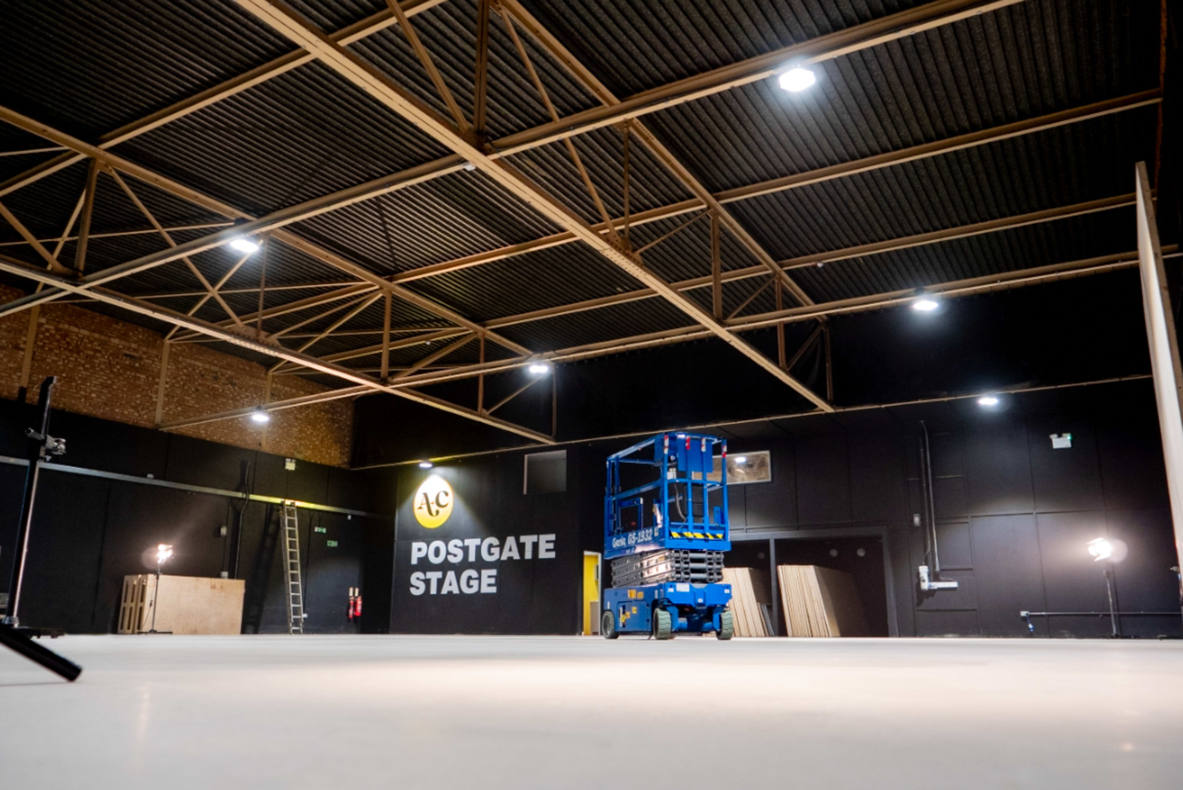 The image shows a spacious industrial stage area with a high ceiling, featuring a blue lift and wooden panels on the floor.