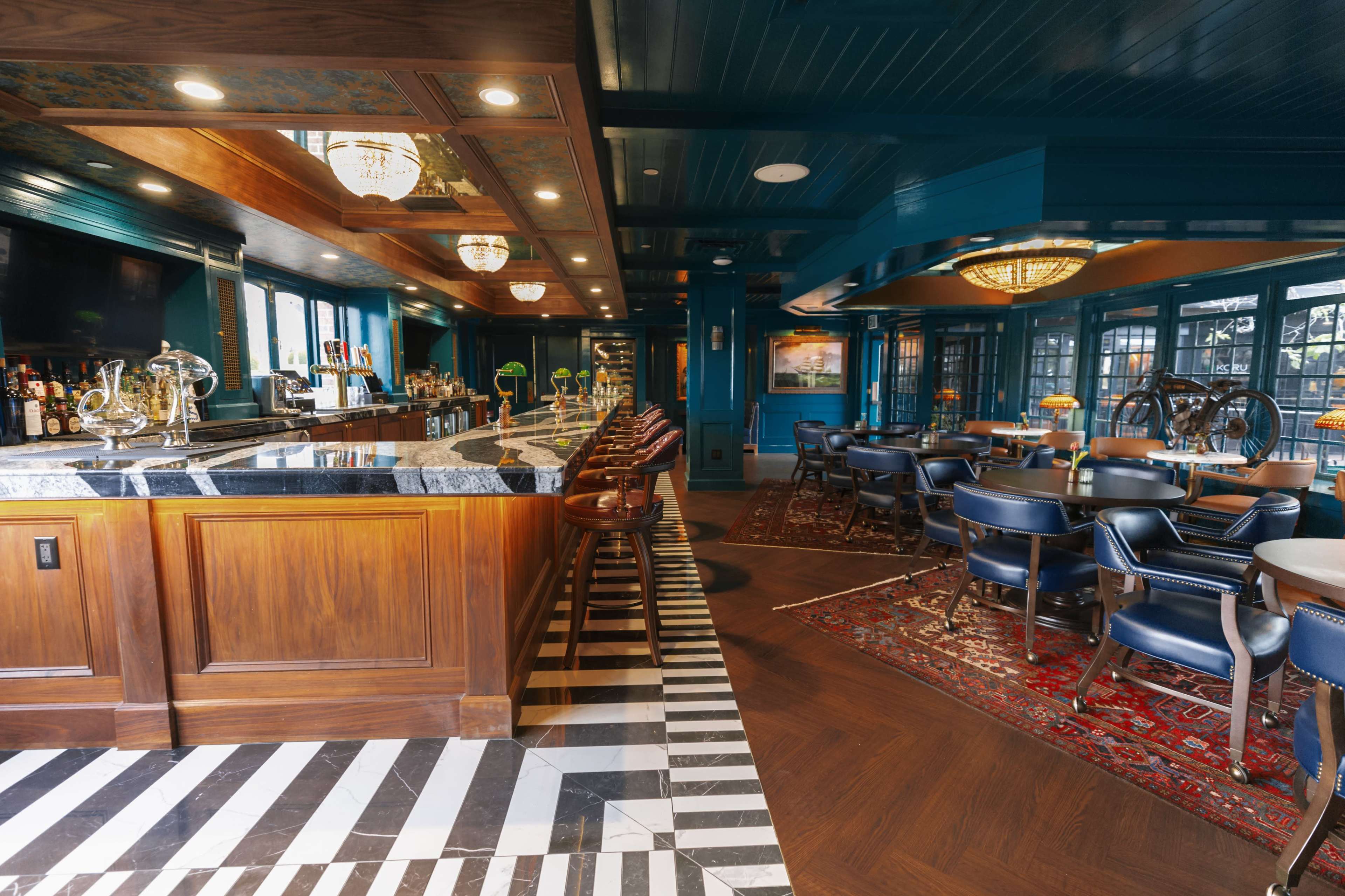 The image shows a stylish bar area with a wooden counter, a variety of liquor bottles, and a patterned floor featuring both dark wood and black-and-white tiles, alongside seating arrangements in a well-lit, elegant setting.