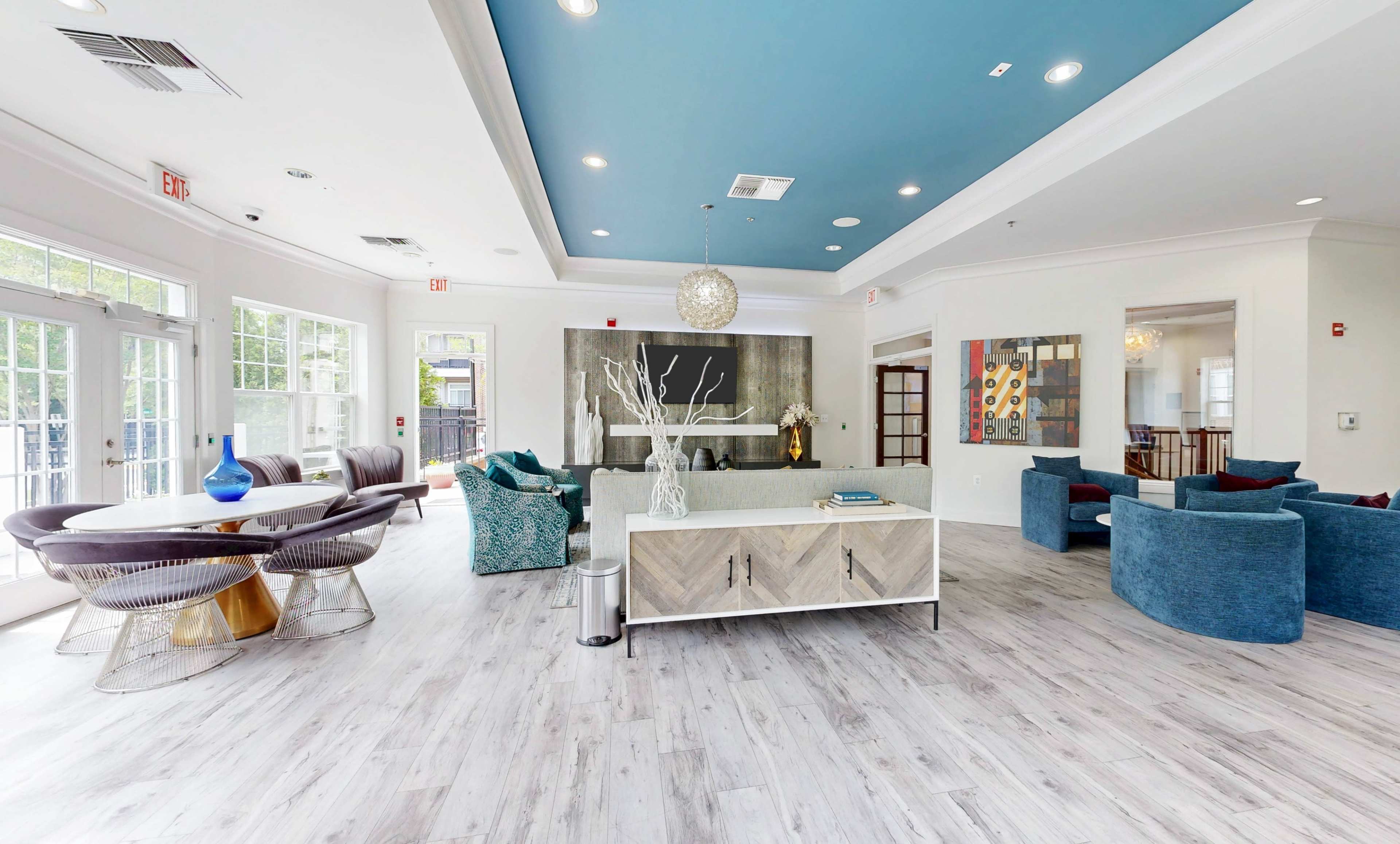 The image shows a modern lounge area featuring a mix of comfortable seating, a round dining table, and a decorative ceiling, all in a well-lit space with large windows.