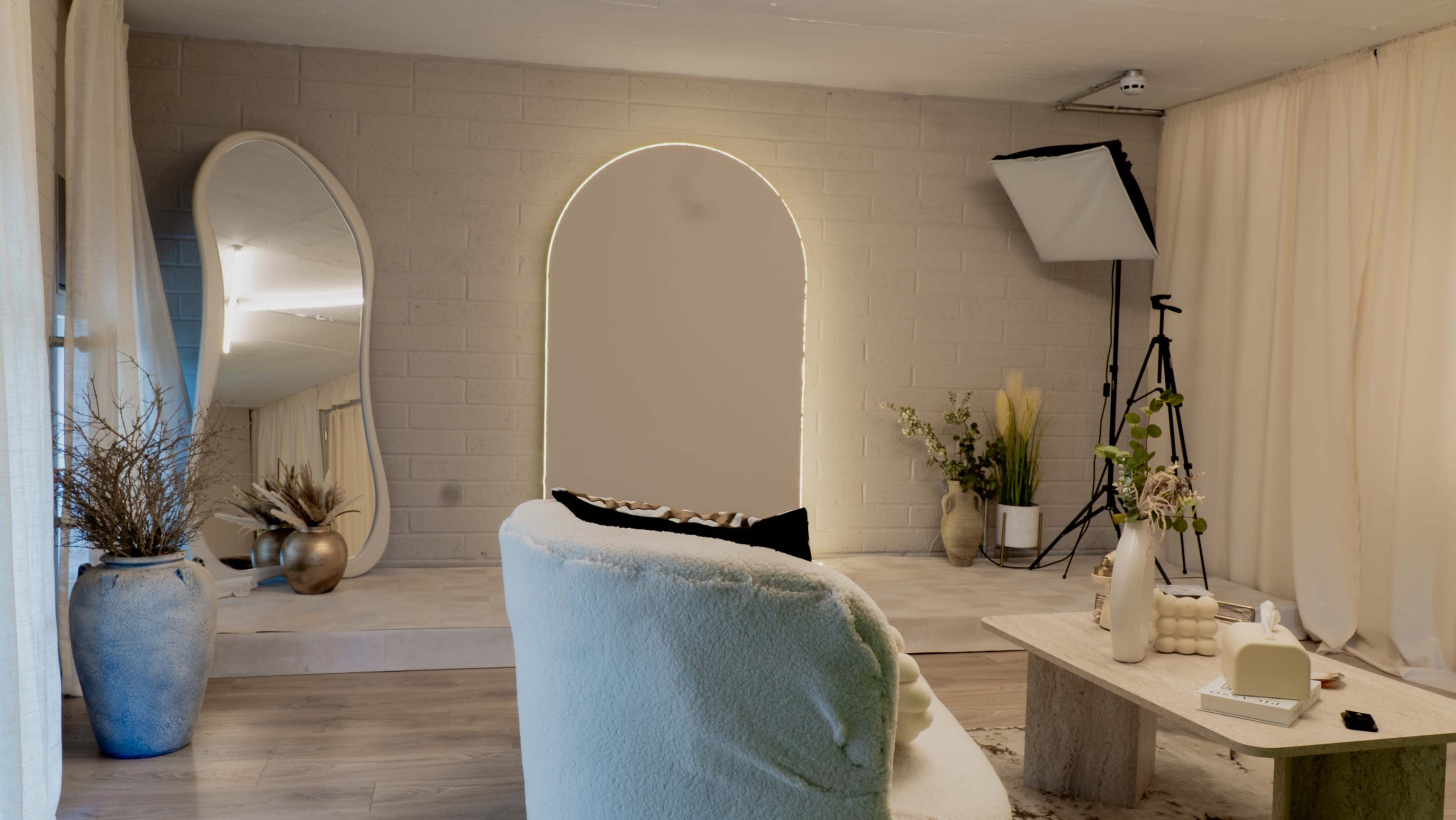 The image shows a minimalist interior space featuring a light-colored wall, a large arched mirror, a sofa, and a wooden table decorated with plants and vases.
