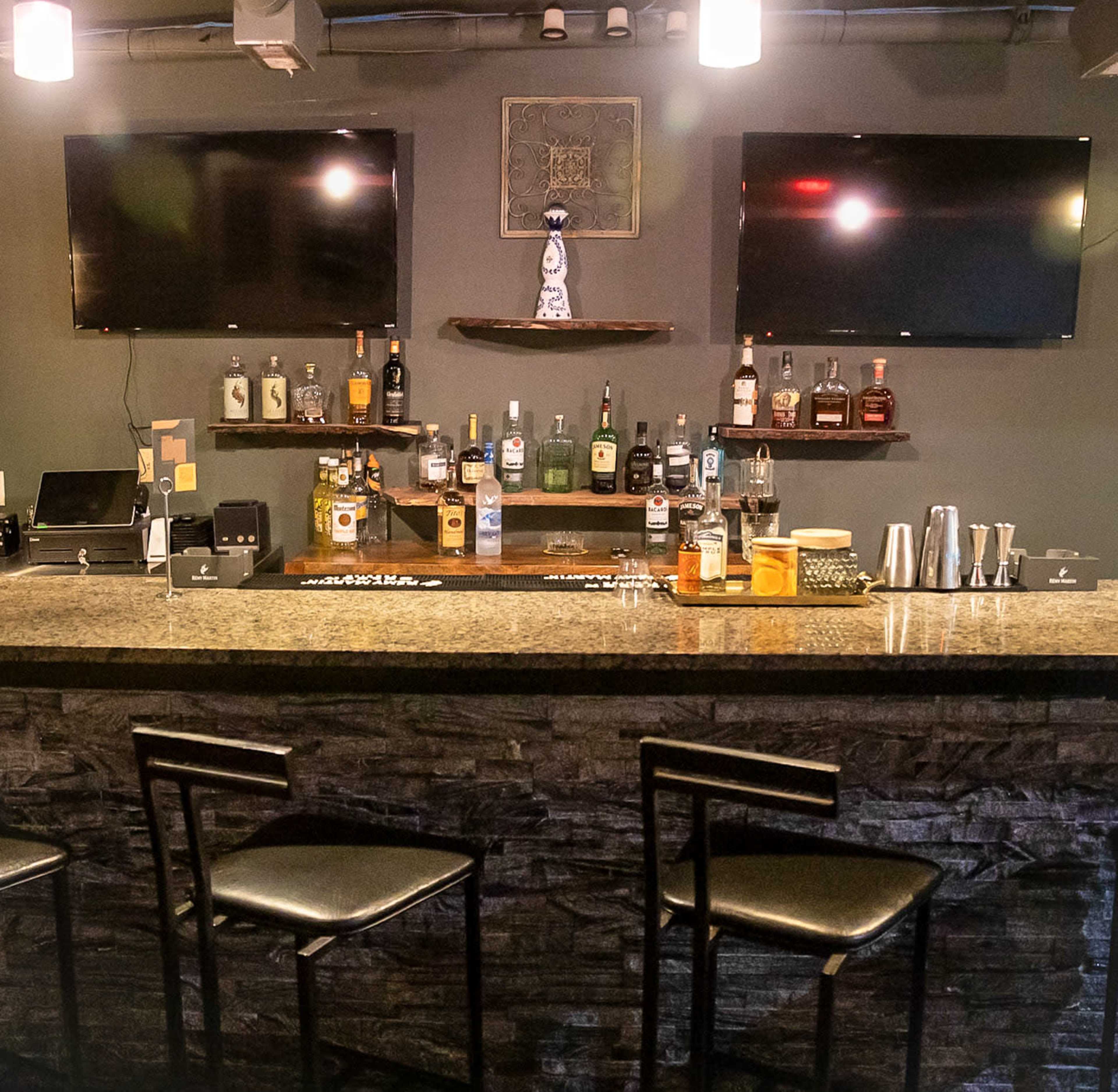 The image shows a modern bar setup featuring a granite countertop, a selection of various liquor bottles on shelves, and two large flat-screen TVs mounted on the wall above.