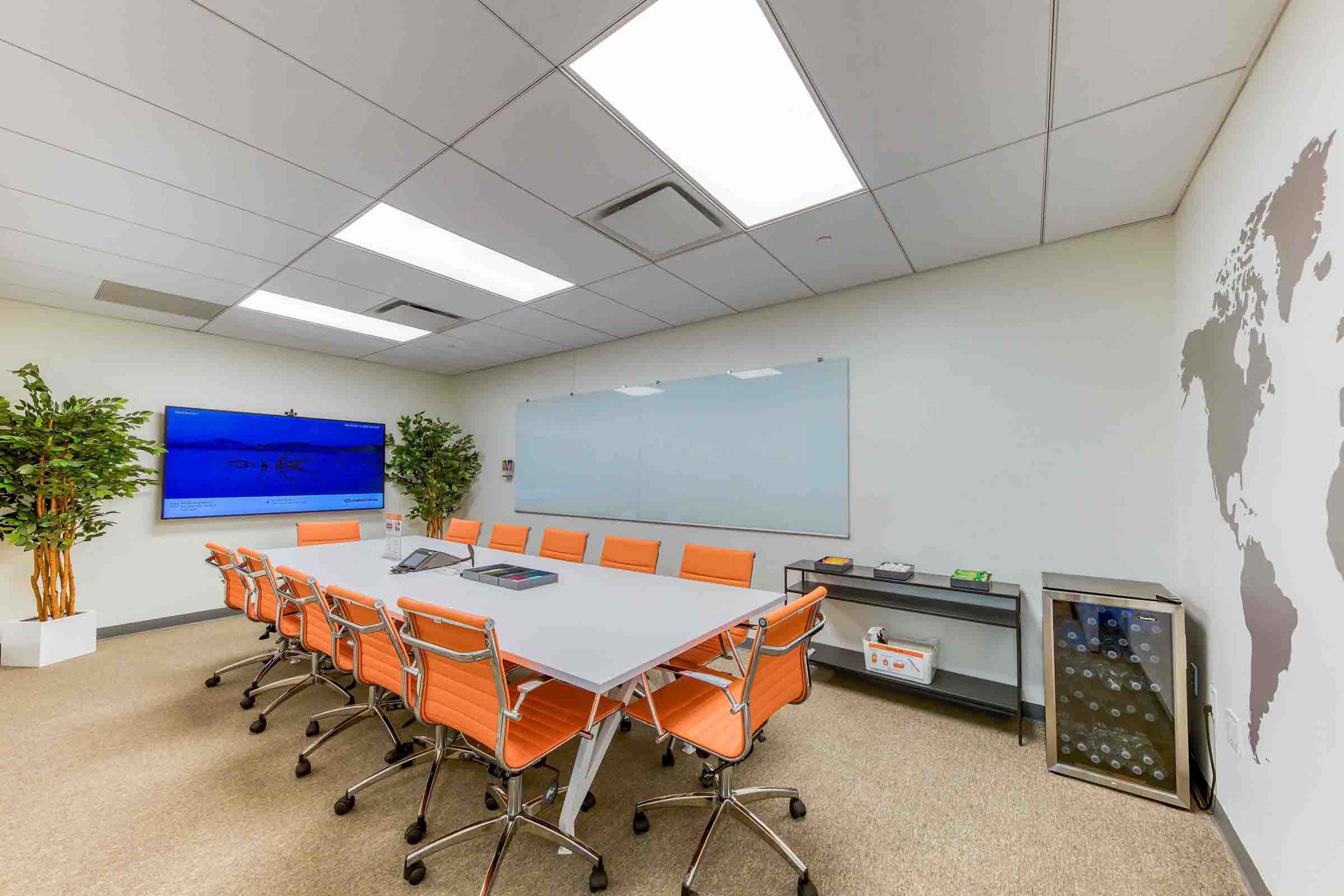 A brightly lit conference room features a large table surrounded by orange chairs, a glass whiteboard, and a display screen on the wall.