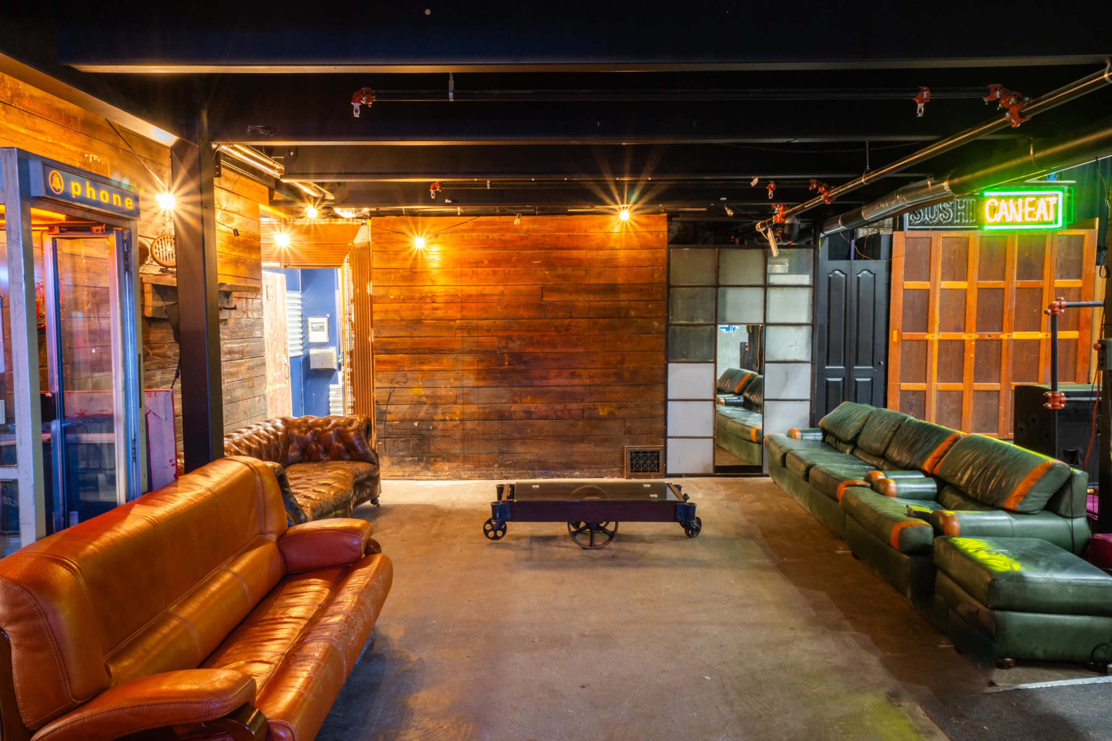 The image shows a sparsely furnished room with wooden walls, featuring two green sofas and a brown leather sofa arranged around a coffee table, with dim lighting and a door leading to another space.