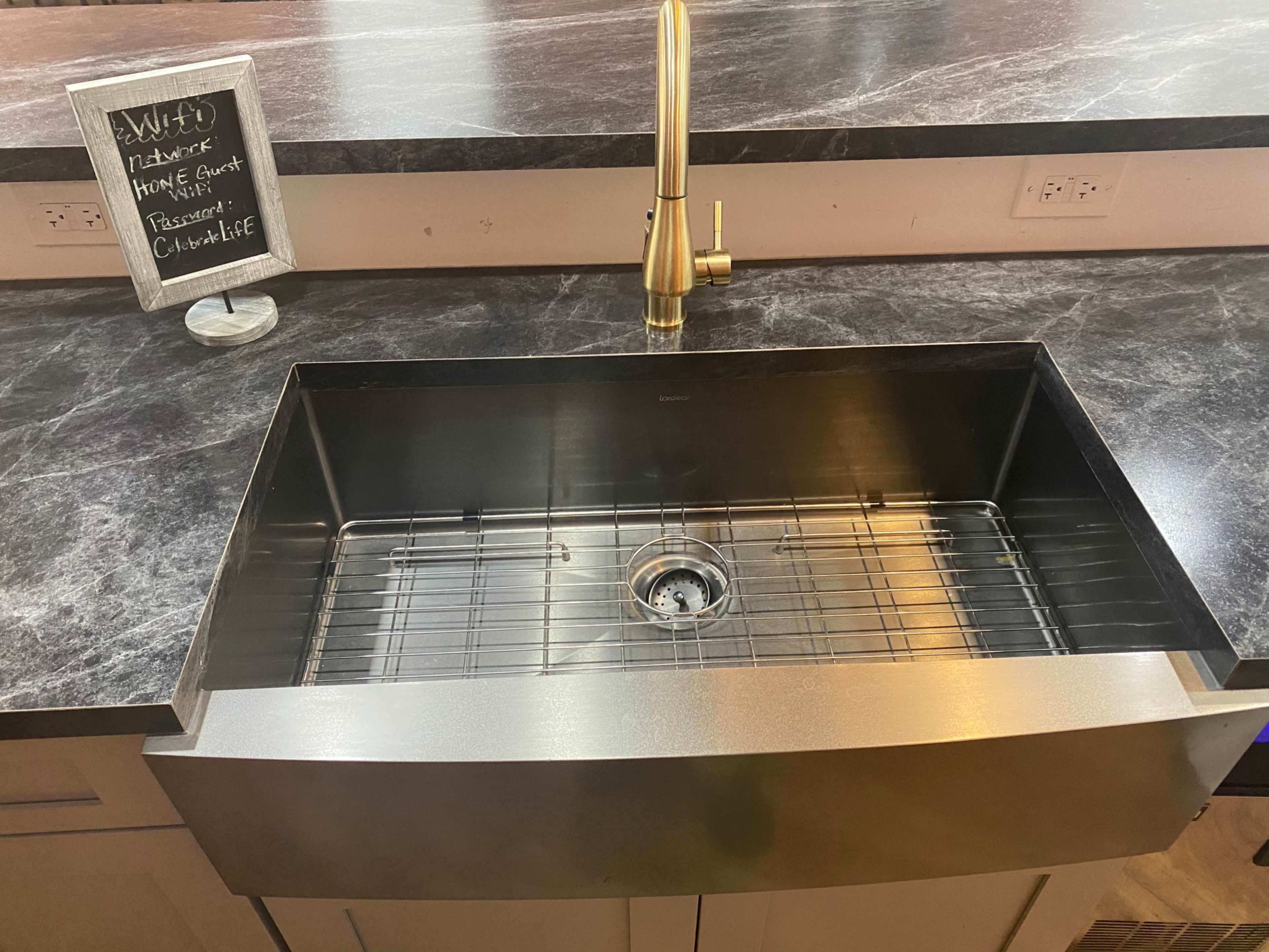 A stainless steel farmhouse sink with a grid insert and a modern gold faucet, set against a dark patterned countertop.