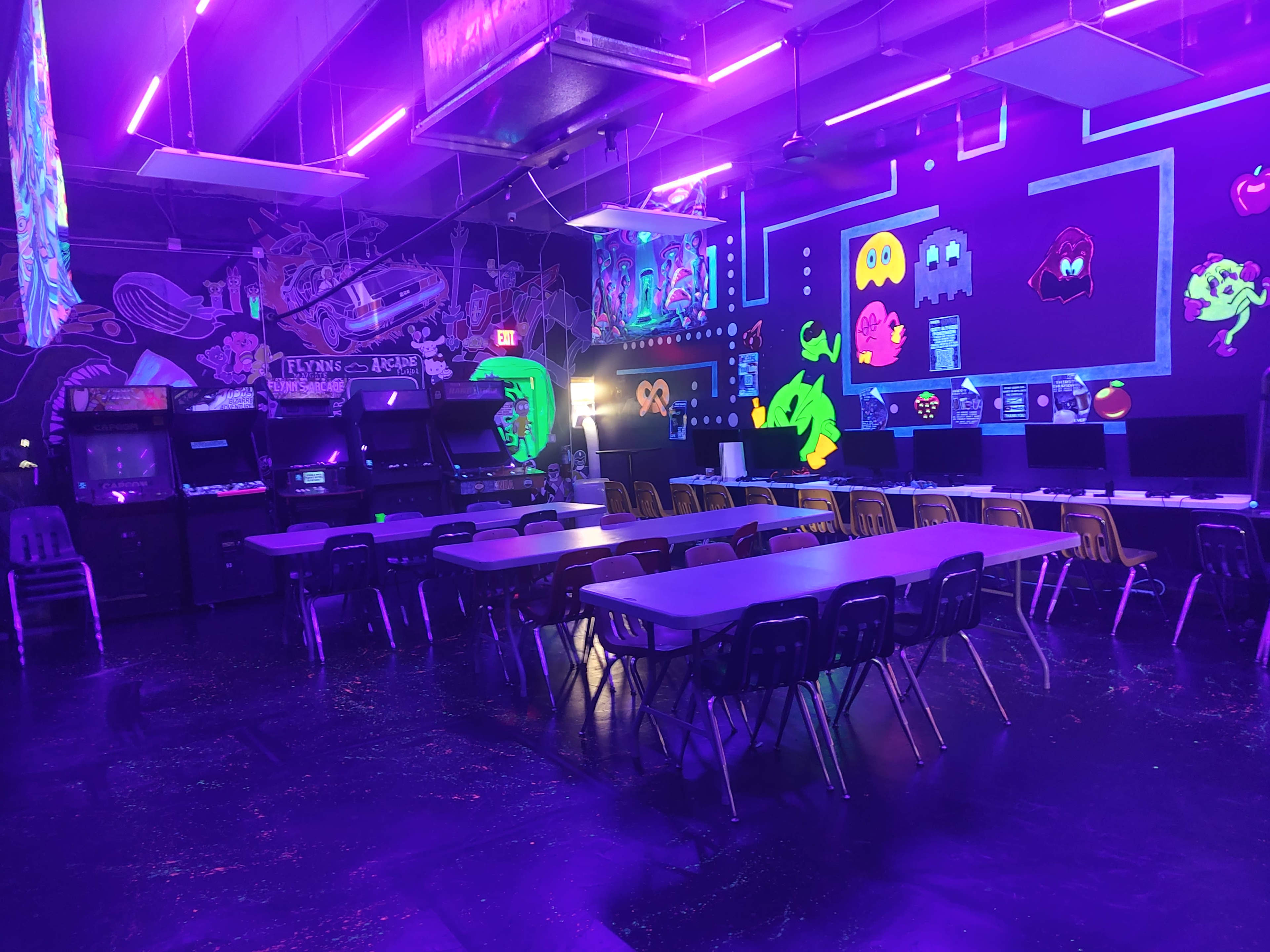 The room features a series of arcade machines lined against one wall, illuminated by purple neon lights, with tables and chairs arranged in the center.