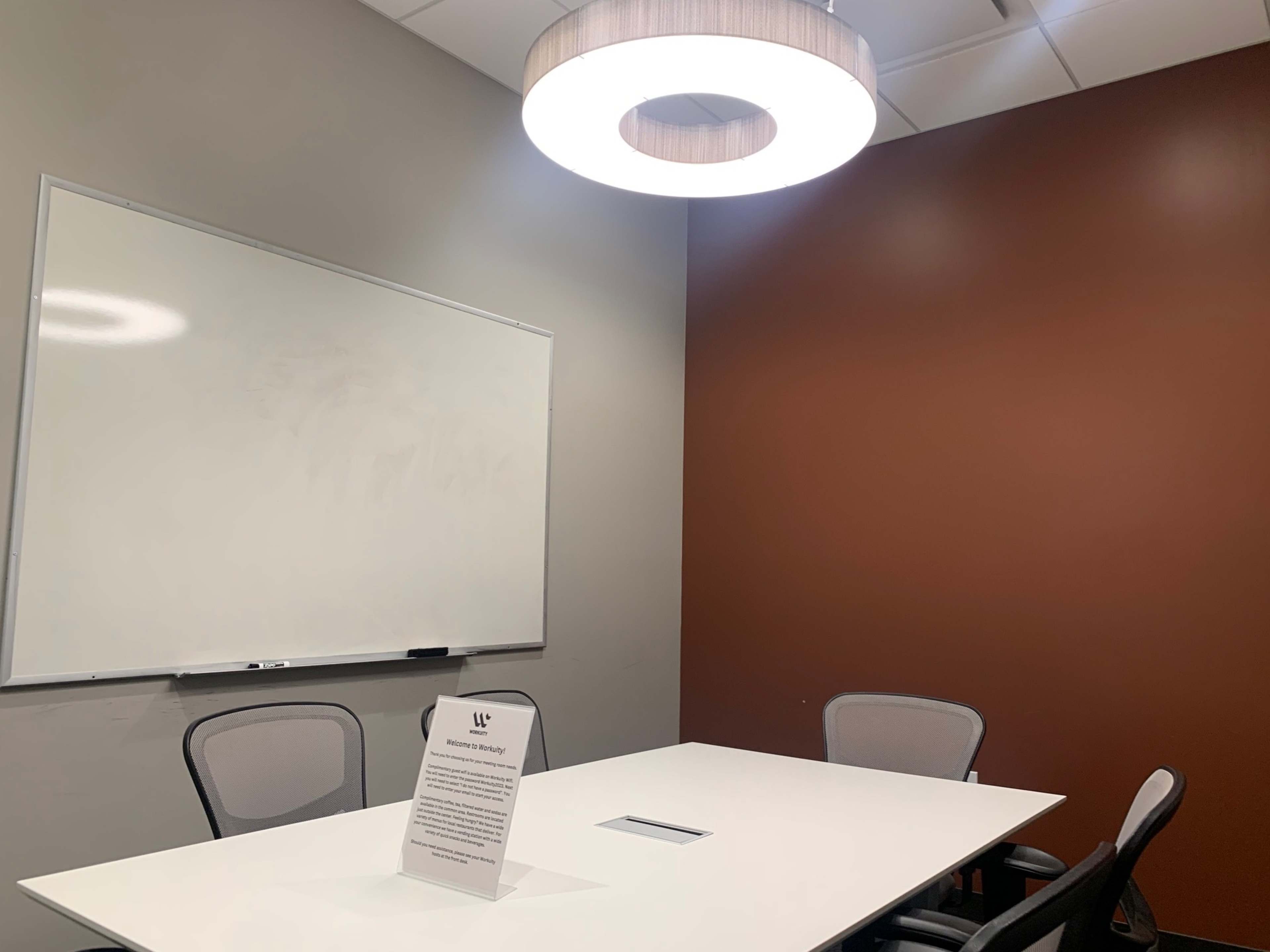 A meeting room features a whiteboard, a contemporary light fixture, a rectangular table, and four chairs, with a sign displayed on the table.