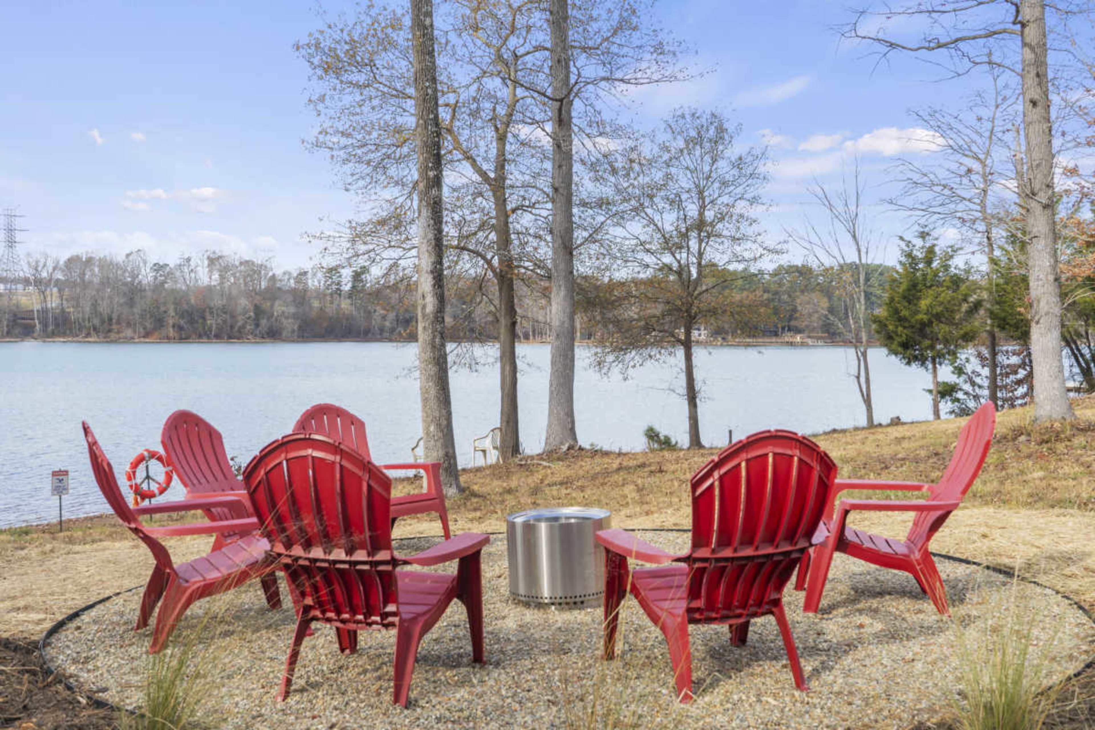 Six red Adirondack chairs are arranged in a circle around a metal fire pit near a lake surrounded by trees.