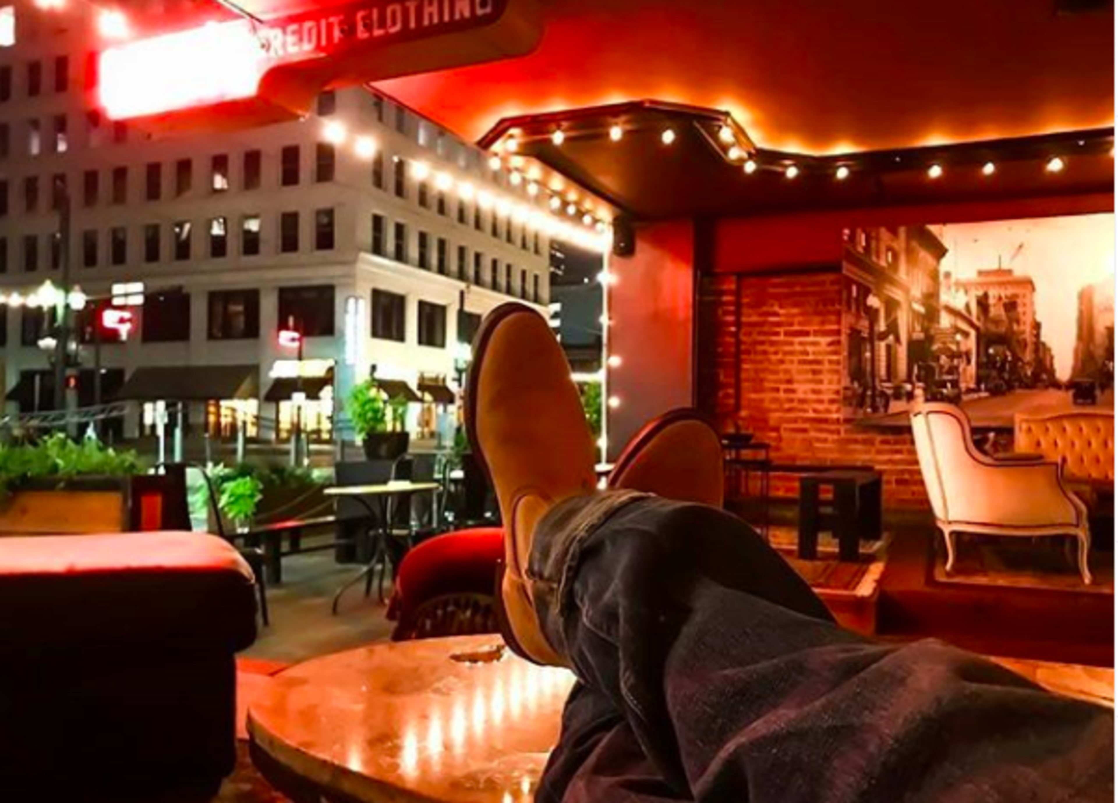 A person relaxes with their feet up on a table in an outdoor seating area, surrounded by twinkling lights and city buildings at night.