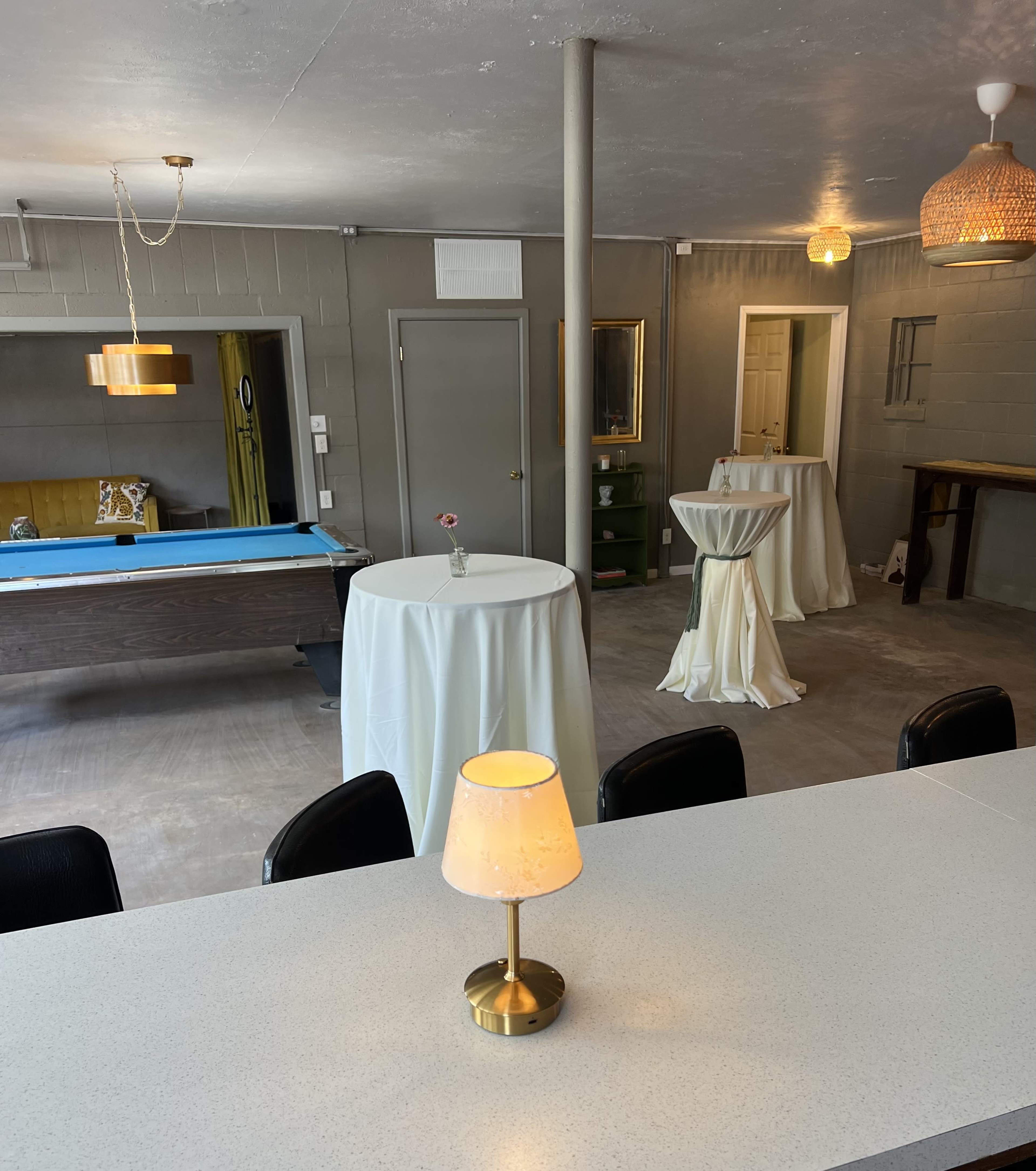 The image shows a spacious interior with a pool table on one side, several round tables with white tablecloths, and decorative lighting fixtures hanging from the ceiling.